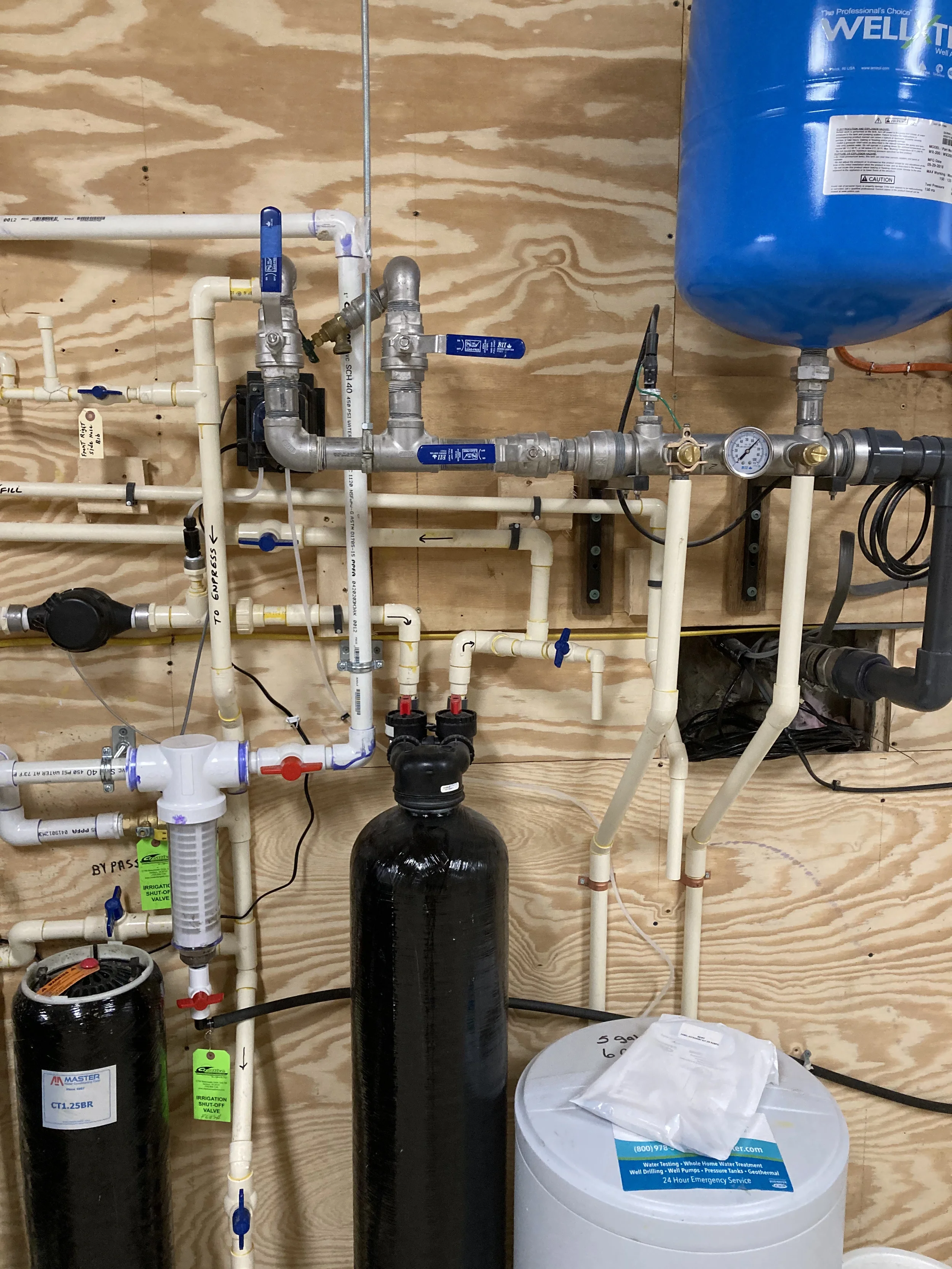 Well pump pressure tank and water treatment manifold installation with valves, gauges, and plumbing mounted on a plywood wall.