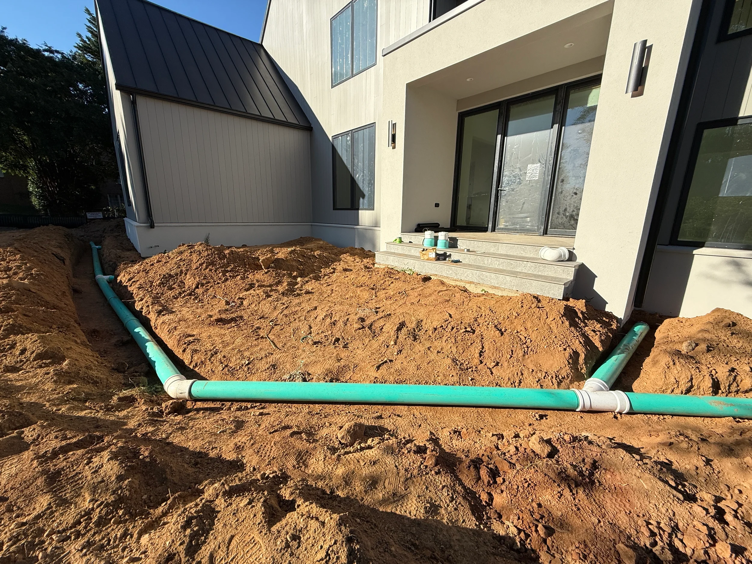 Perimeter stormwater pipe routing around a rear patio foundation using green PVC drain lines during residential site drainage construction.