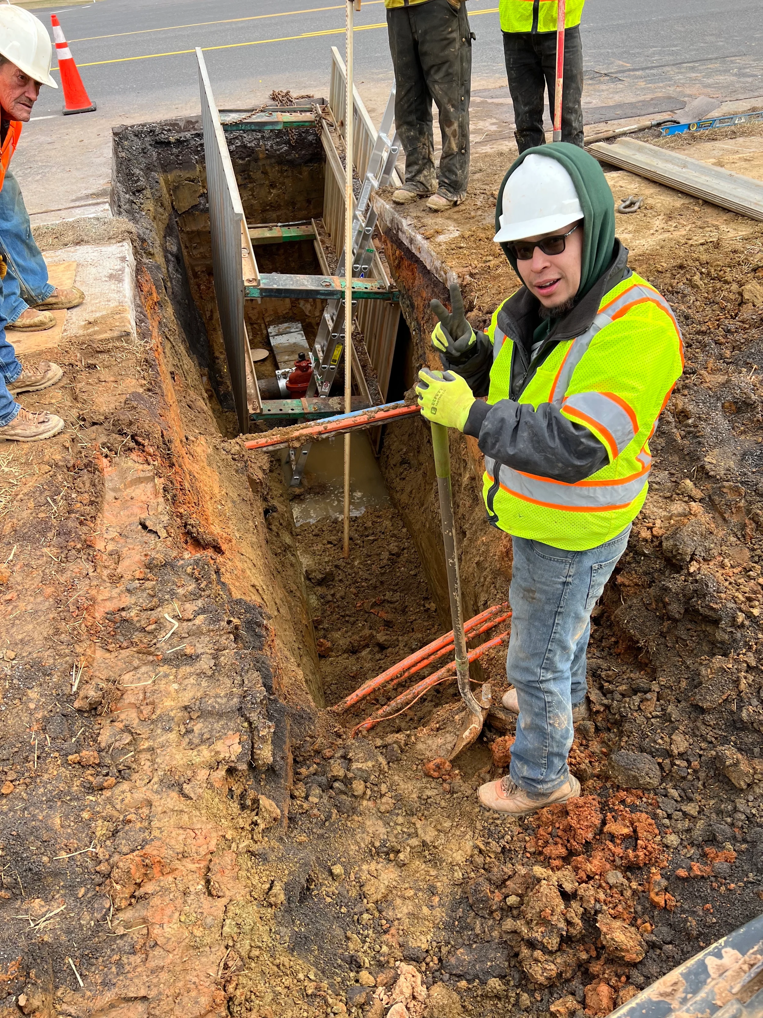 Roadside water main repair with crew working around a shored trench, valve connection, ladder access, and excavation safety controls.