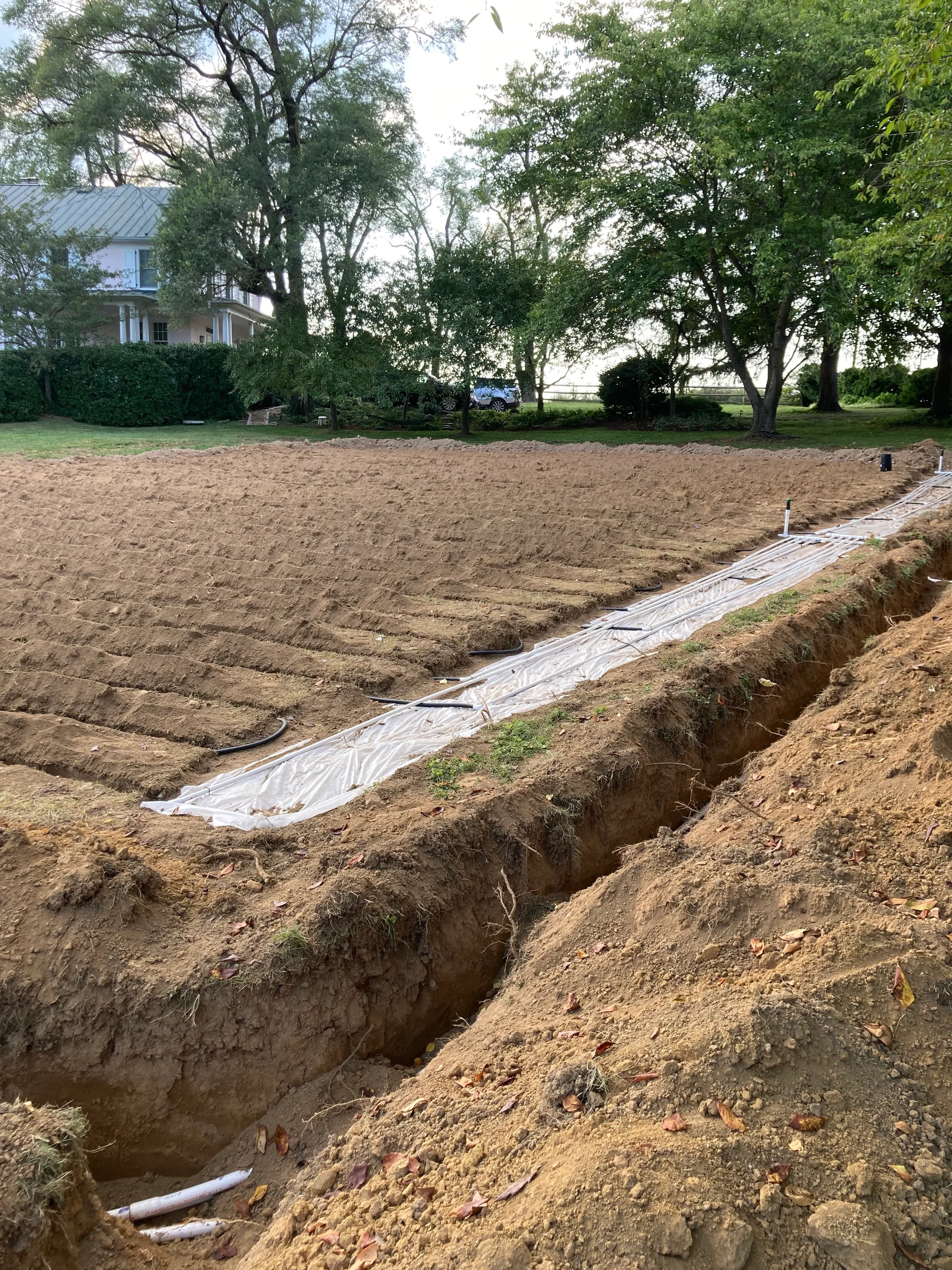 Septic supply trench lined and piped across a graded drainfield in front of a farmhouse during a rural Northern Virginia septic installation.