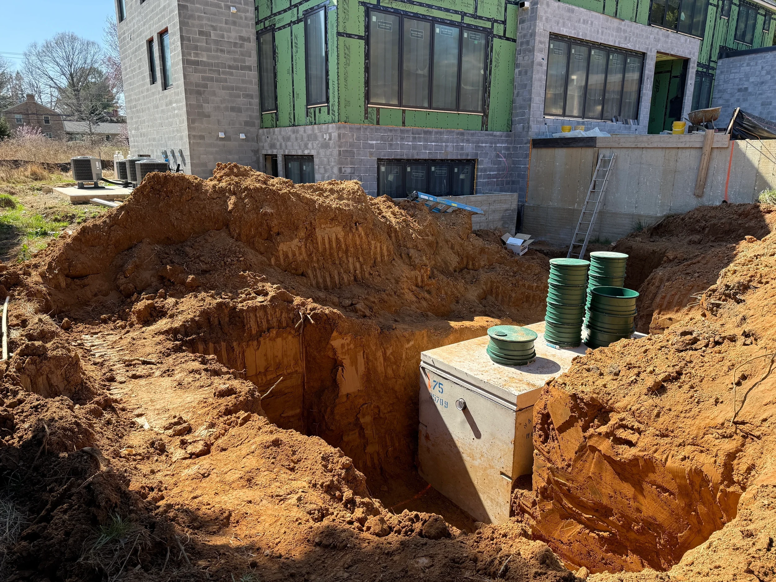 Precast septic tank set in its excavation with riser lids beside a home under construction, with AC units and an access ladder visible on the Virginia jobsite.