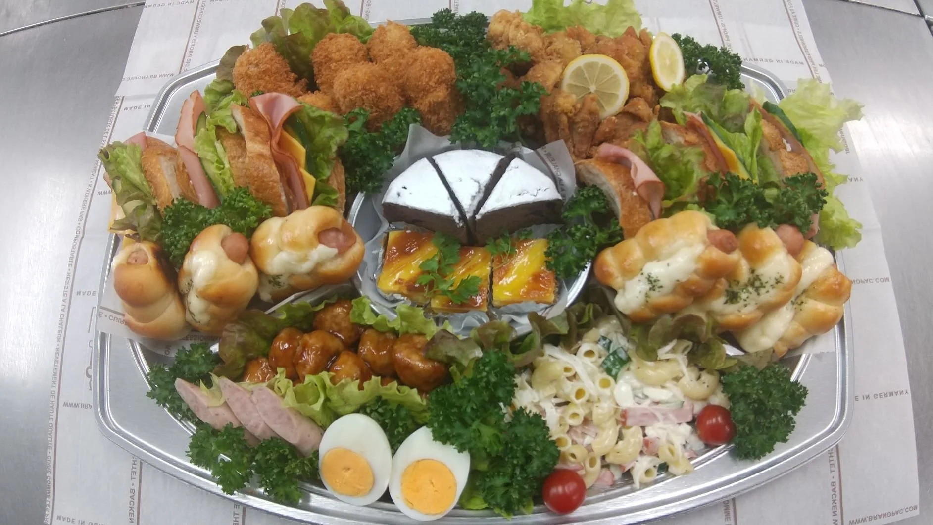 さまざまな種類の惣菜パン、フライドチキン、コロッケ、サラダ、チョコケーキ、ゆで卵などが盛り付けられた大きなトレイ