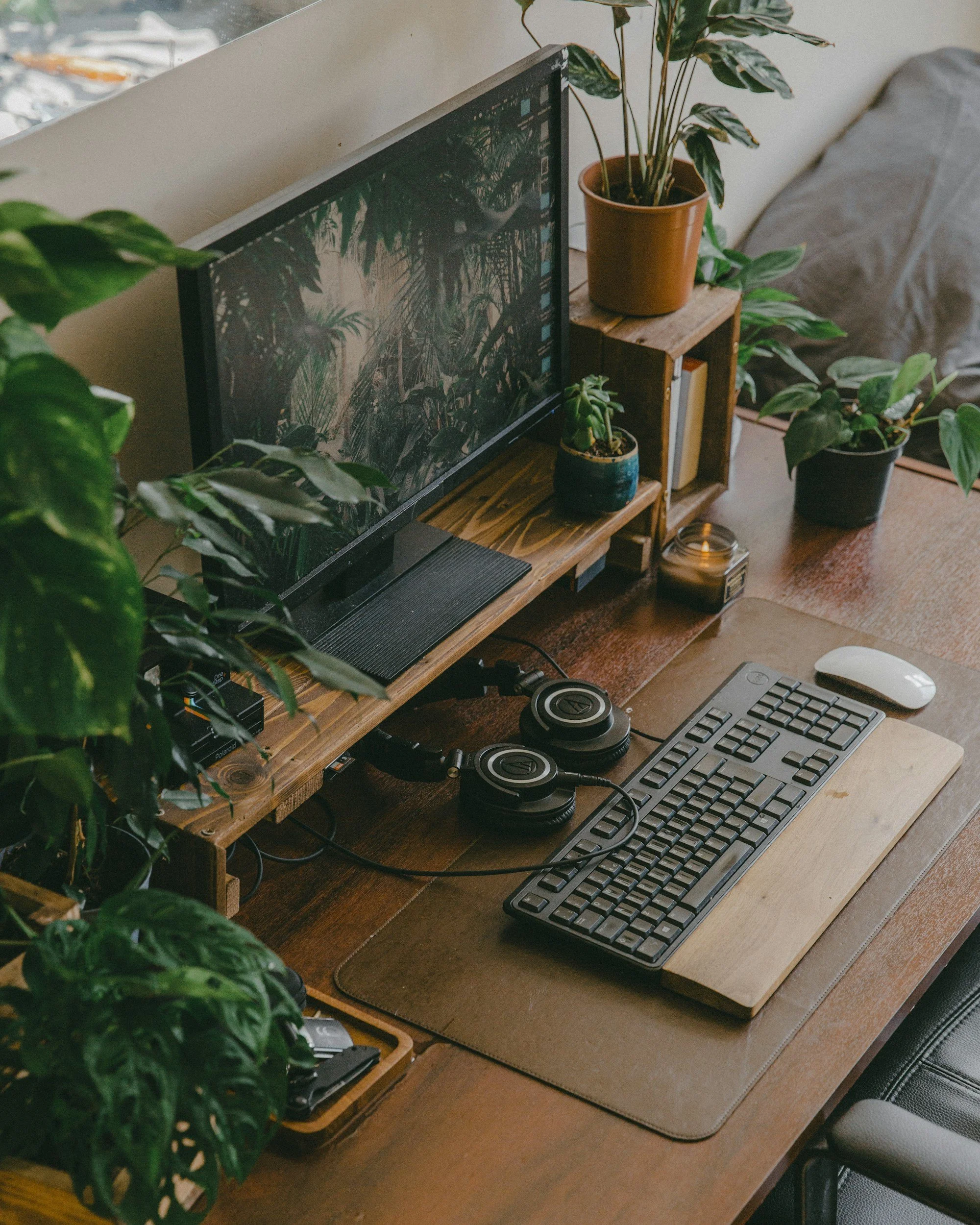 A workspace with a wooden desk holding a computer monitor, keyboard, mouse, and headphones. Green potted plants are on a small wooden shelf and the desk. There is a blanket on a chair and a window in the background.