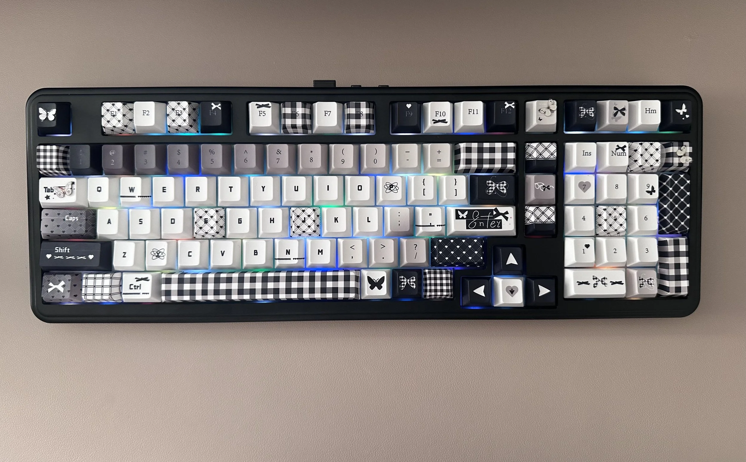 Top view of a computer keyboard with black, white, and checkered keycaps, including butterfly and floral designs, placed on a beige surface.
