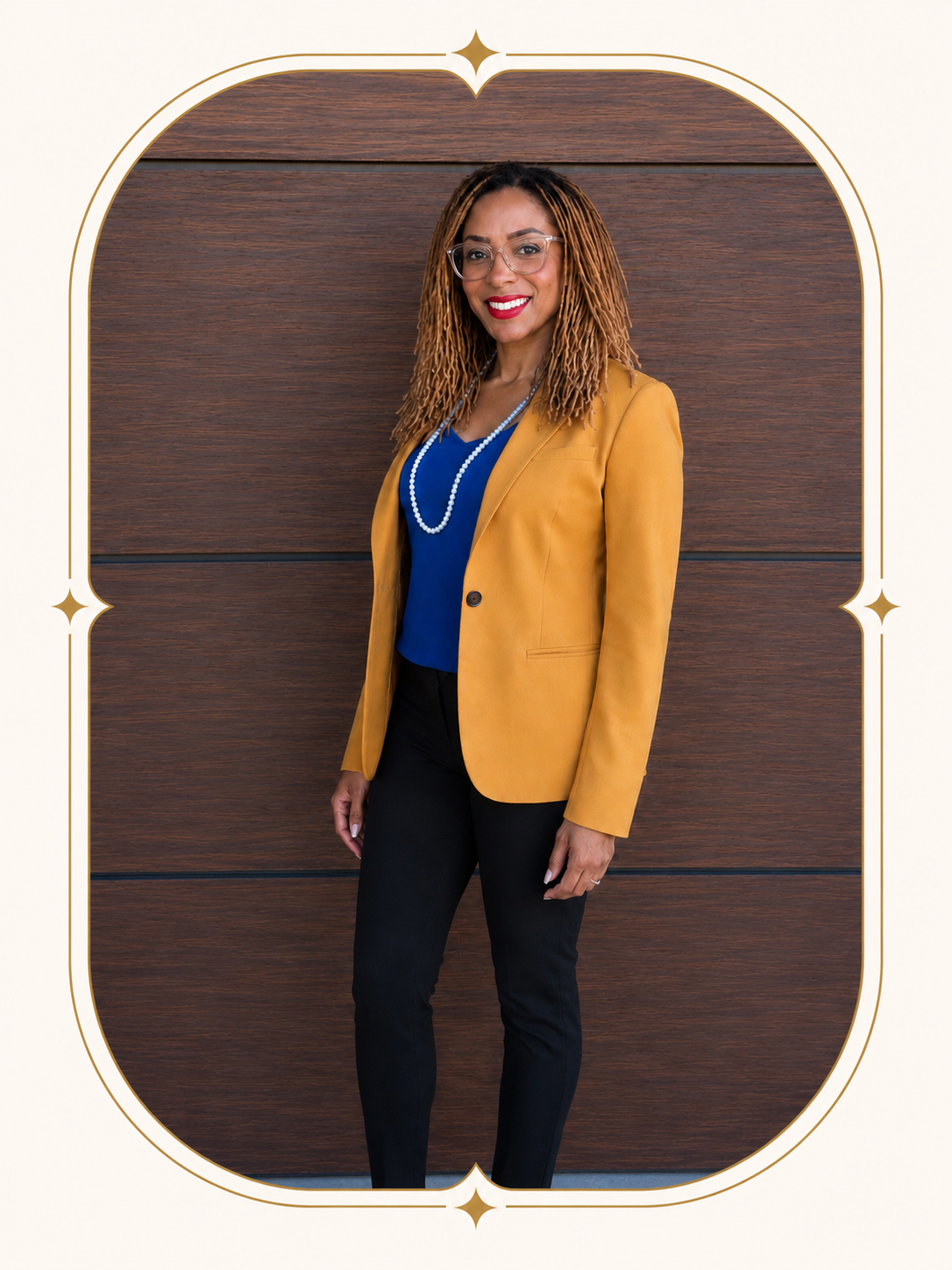 A woman with shoulder-length curly hair wearing glasses, a mustard yellow blazer, a blue top, black pants, and a pearl necklace standing against a wooden wall.