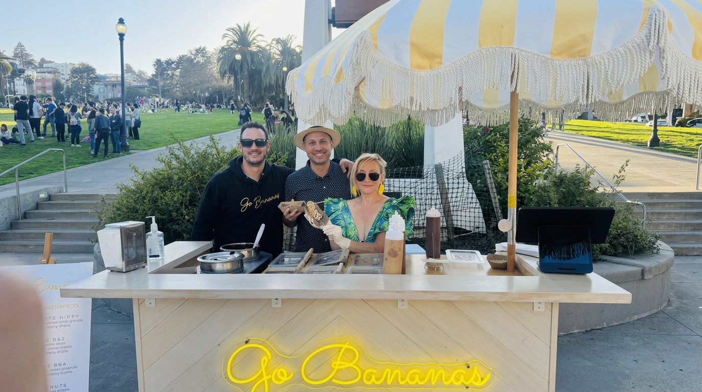 A food stand with a yellow neon sign reading 'Go Bananas' in front of a park on a sunny day. Three people are behind the stand smiling, with a man wearing sunglasses and a woman with yellow earrings and dark sunglasses in the center. The stand has various items including a large bottle, a tablet, and containers for food preparation.