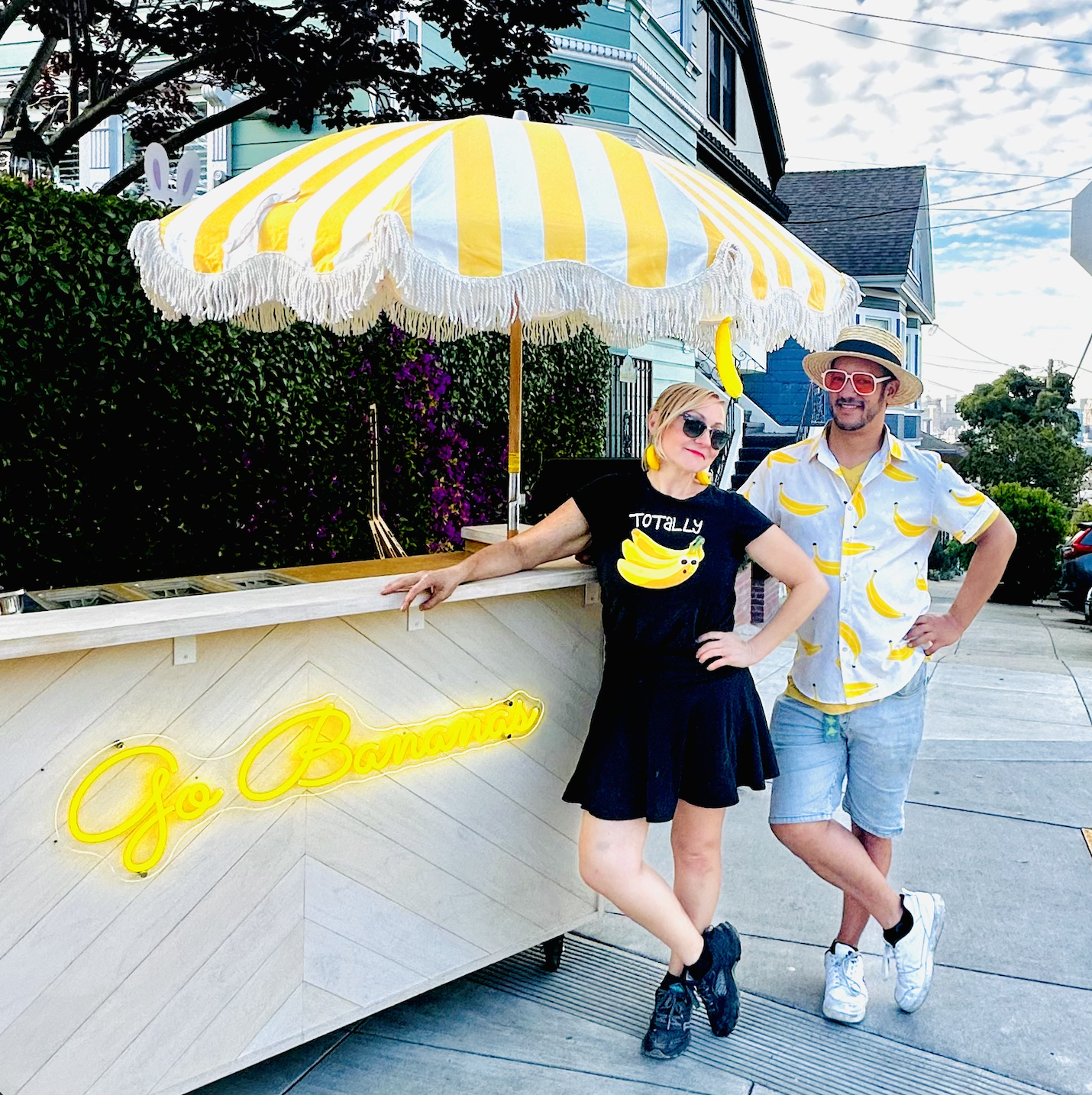 Two people standing outside a banana stand with a yellow and white striped umbrella. The woman is wearing a black shirt with a banana graphic and the words 'Totally Bananas.' The man is wearing a short-sleeved shirt with banana prints, a straw hat, and sunglasses. The stand has a neon sign that says 'Go Bananas'.
