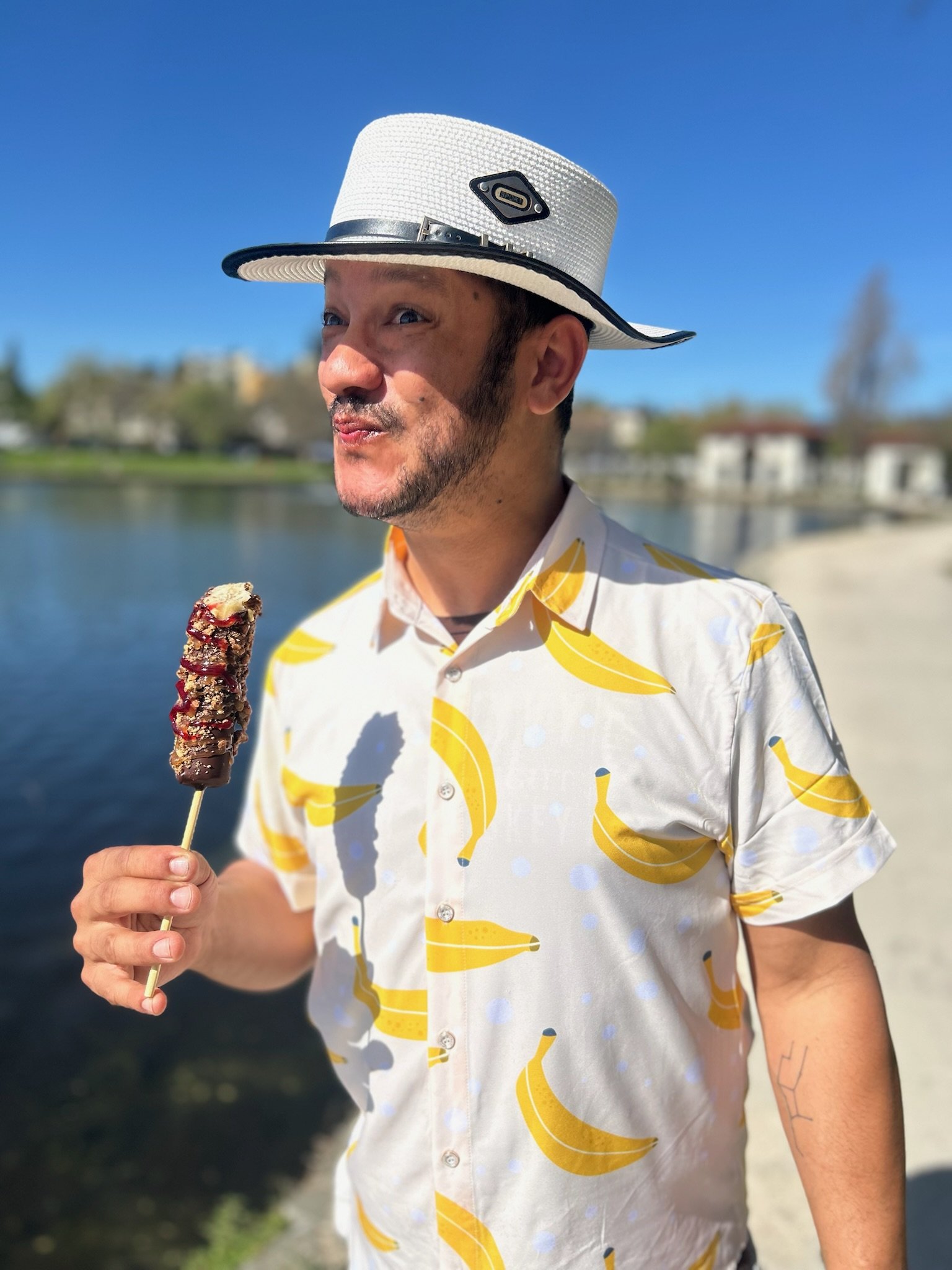 A man wearing a white hat and a yellow-patterned shirt is holding a chocolate-dipped frozen banana standing outdoors by a lake.