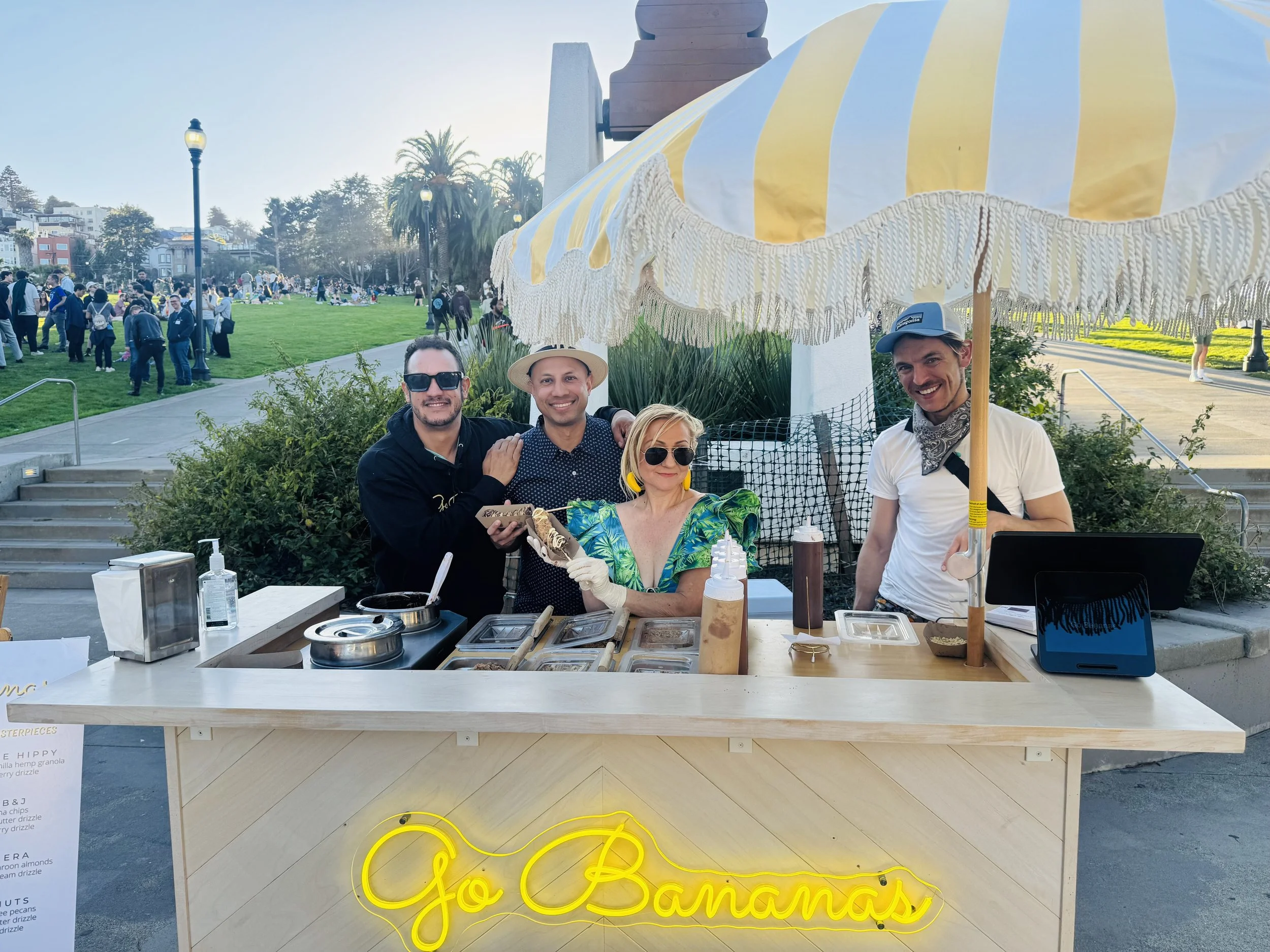 Four people posing behind a counter at an outdoor food stand. The stand has a yellow neon sign reading 'Go Bananas'. There are various toppings on the counter. The background shows a park with many people, some sitting on the grass, and some walking or standing.