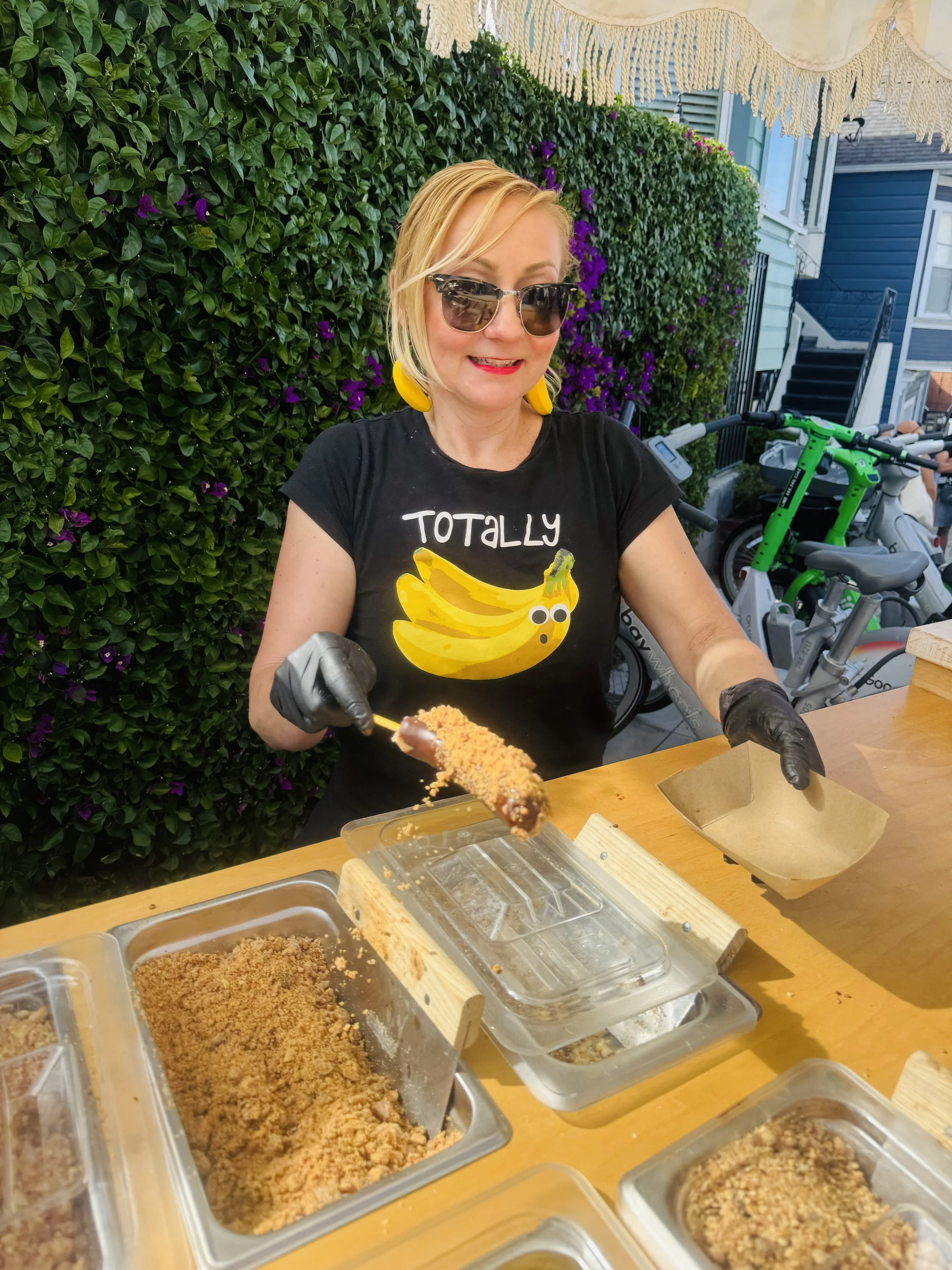 A woman wearing sunglasses and yellow earrings is preparing a frozen chocolate dipped banana, with containers of toppings in front of her. There are purple flowers and greenery behind her, and bicycles are parked nearby.