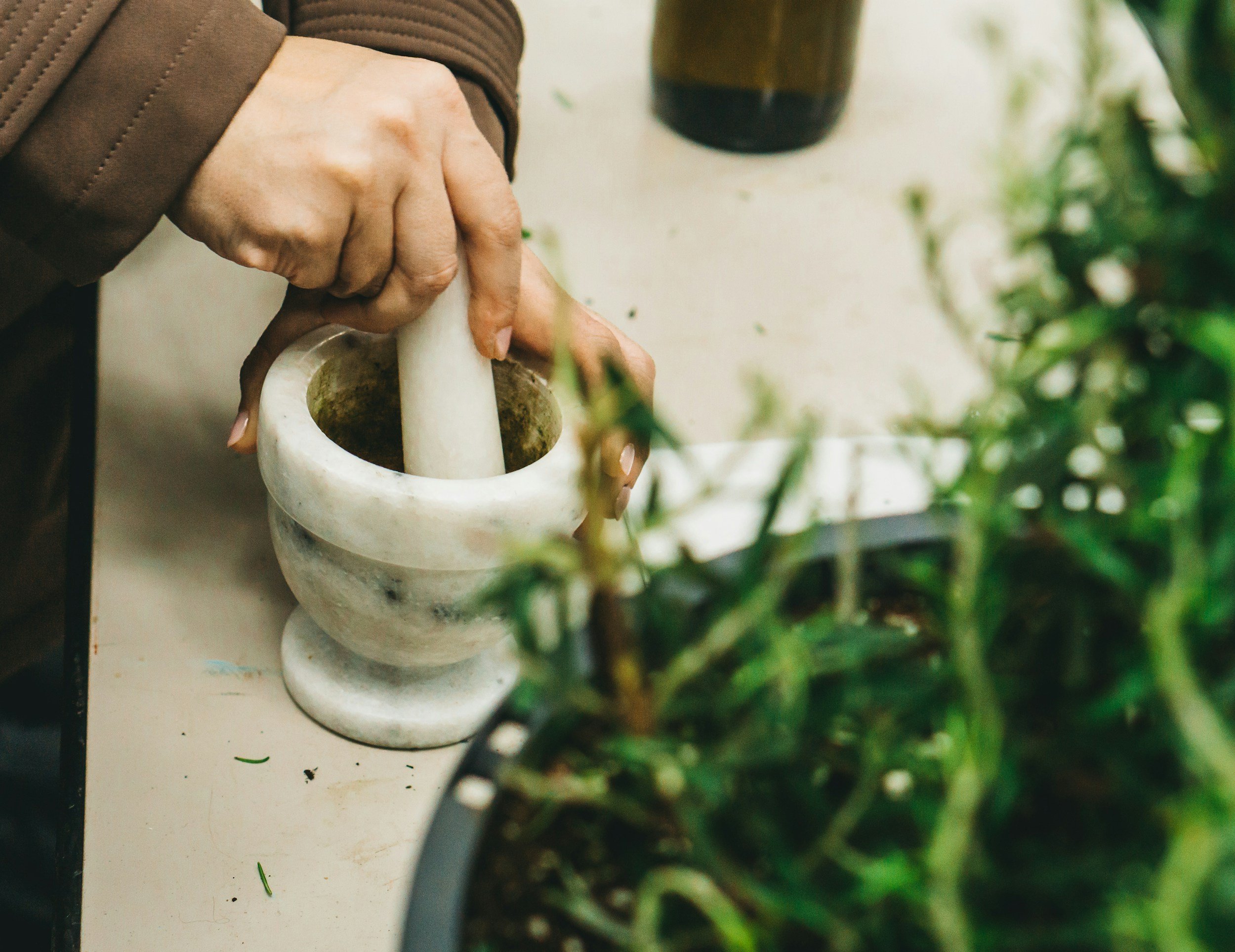 Person in a brown jacket grinding spices or herbs using a mortar and pestle on a white surface, with a dark glass jar nearby and green plants in the background.