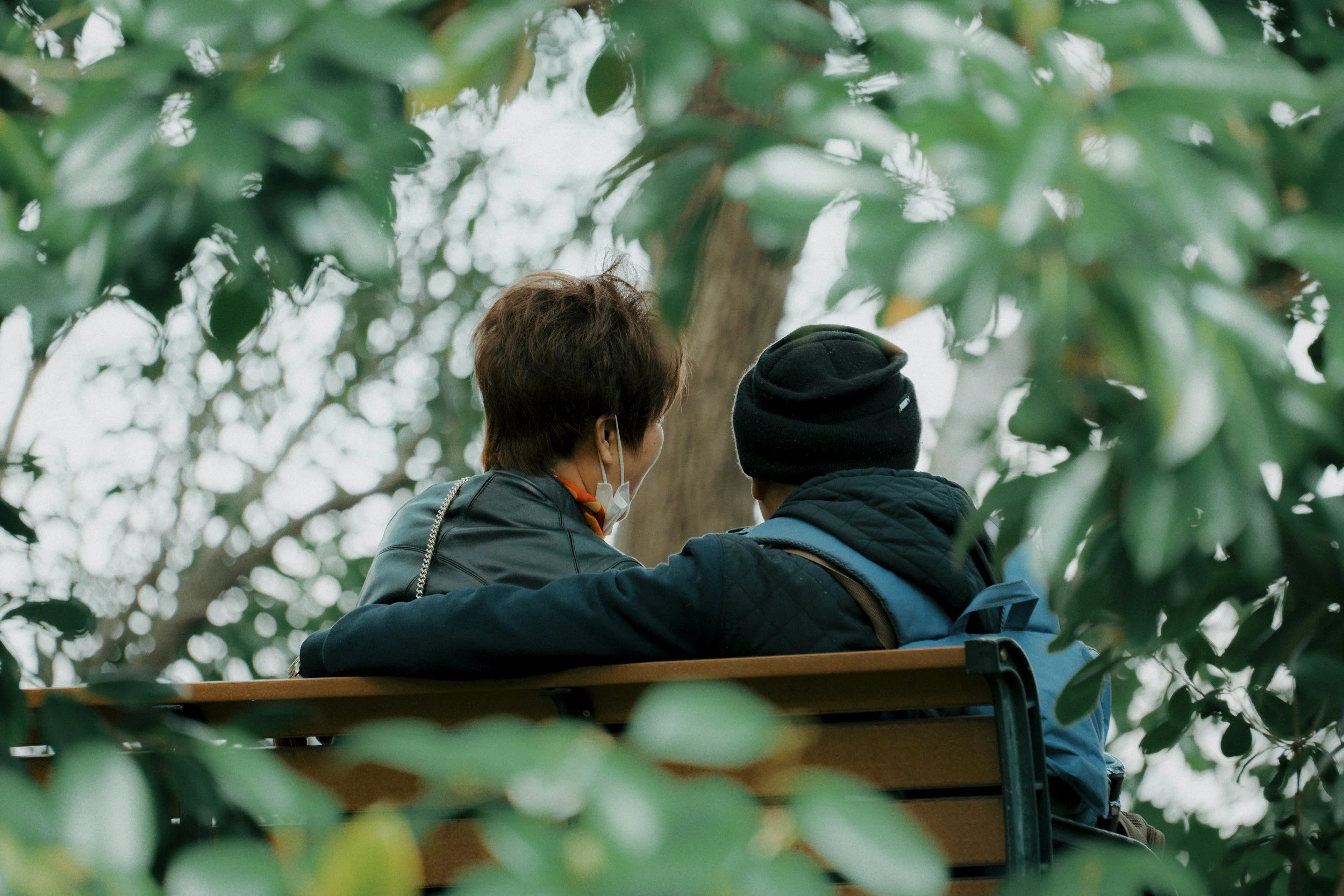 Two people sitting closely together outdoors on a bench, surrounded by green foliage, with one wearing a face mask and the other wearing a black beanie.