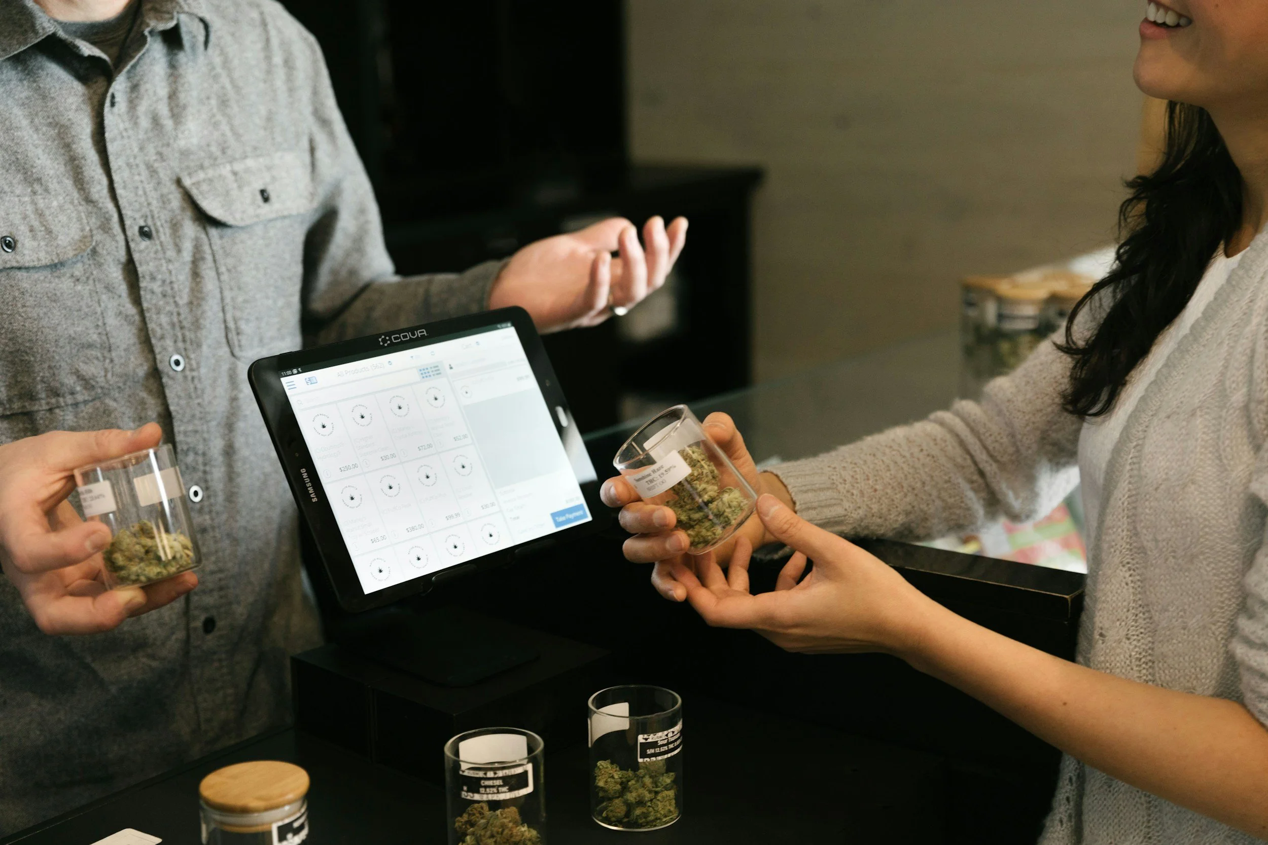 People at a store counter talking about cannabis products. One person is holding a small jar filled with cannabis buds, while another is adjusting an electronic device displaying product prices and details.