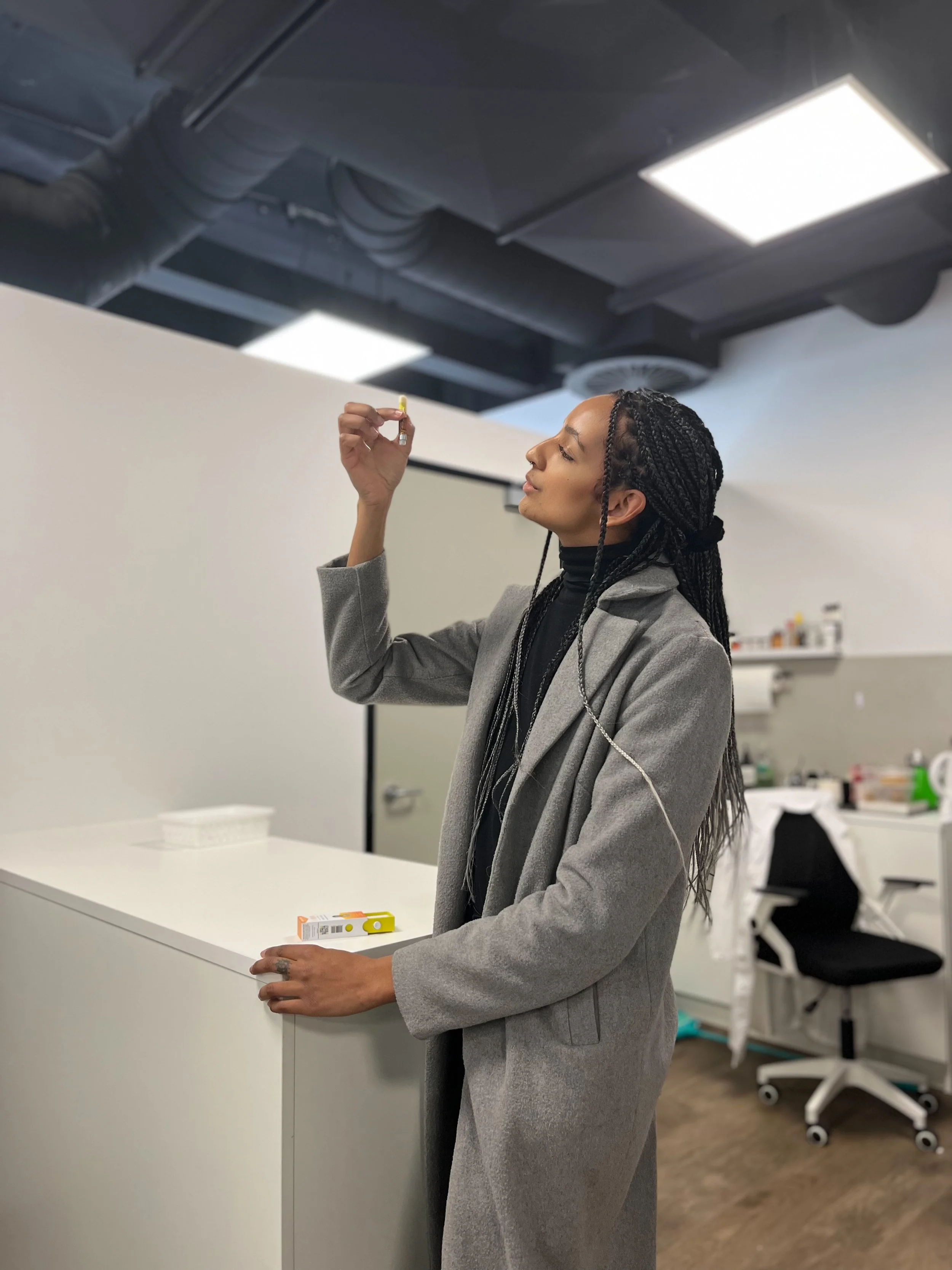 A woman with braided hair and a gray coat holding a small box, standing in a modern office or lab space with a white counter and office chairs in the background.