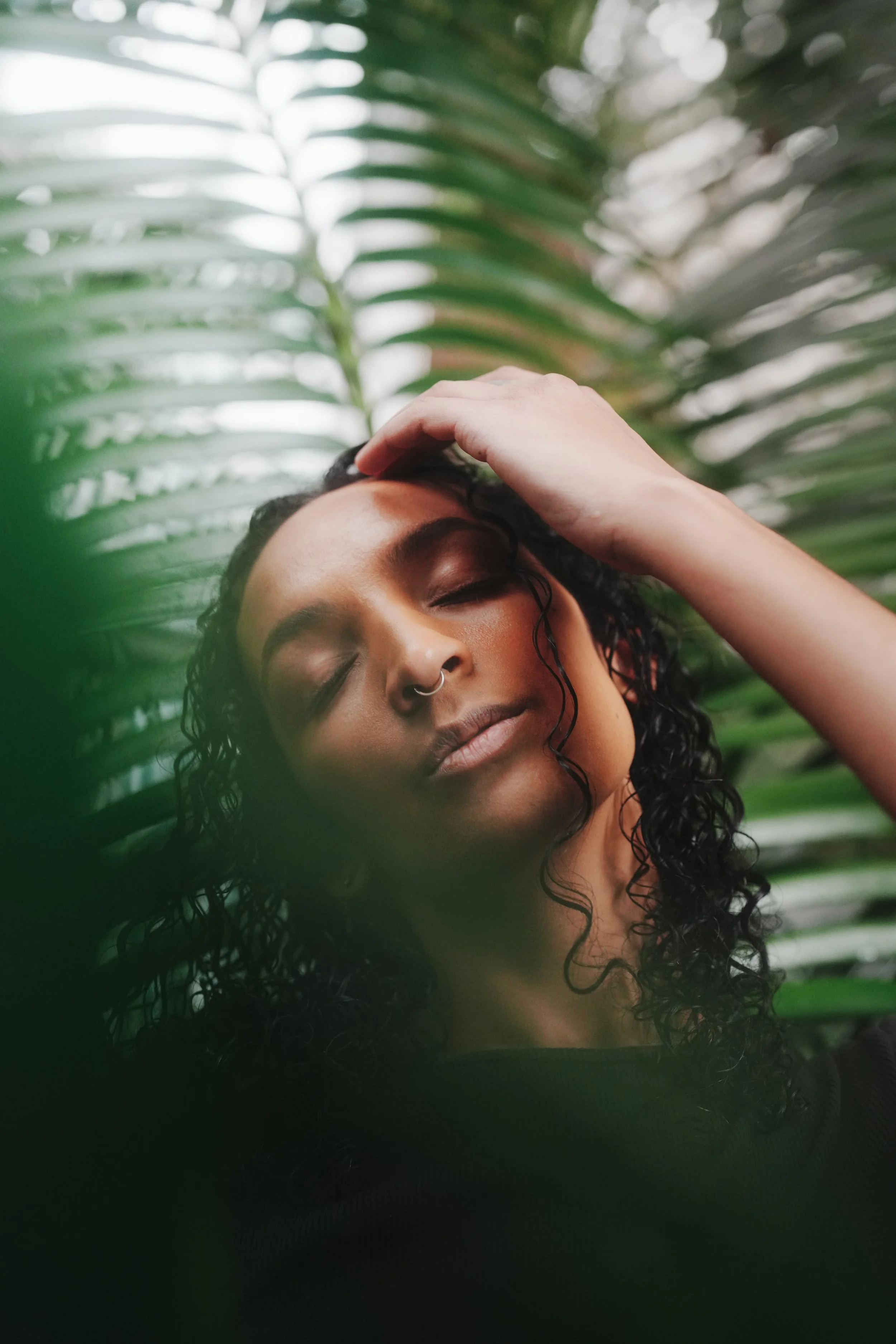 A woman with curly hair and a septum piercing resting her hand on her forehead, surrounded by large green palm leaves.