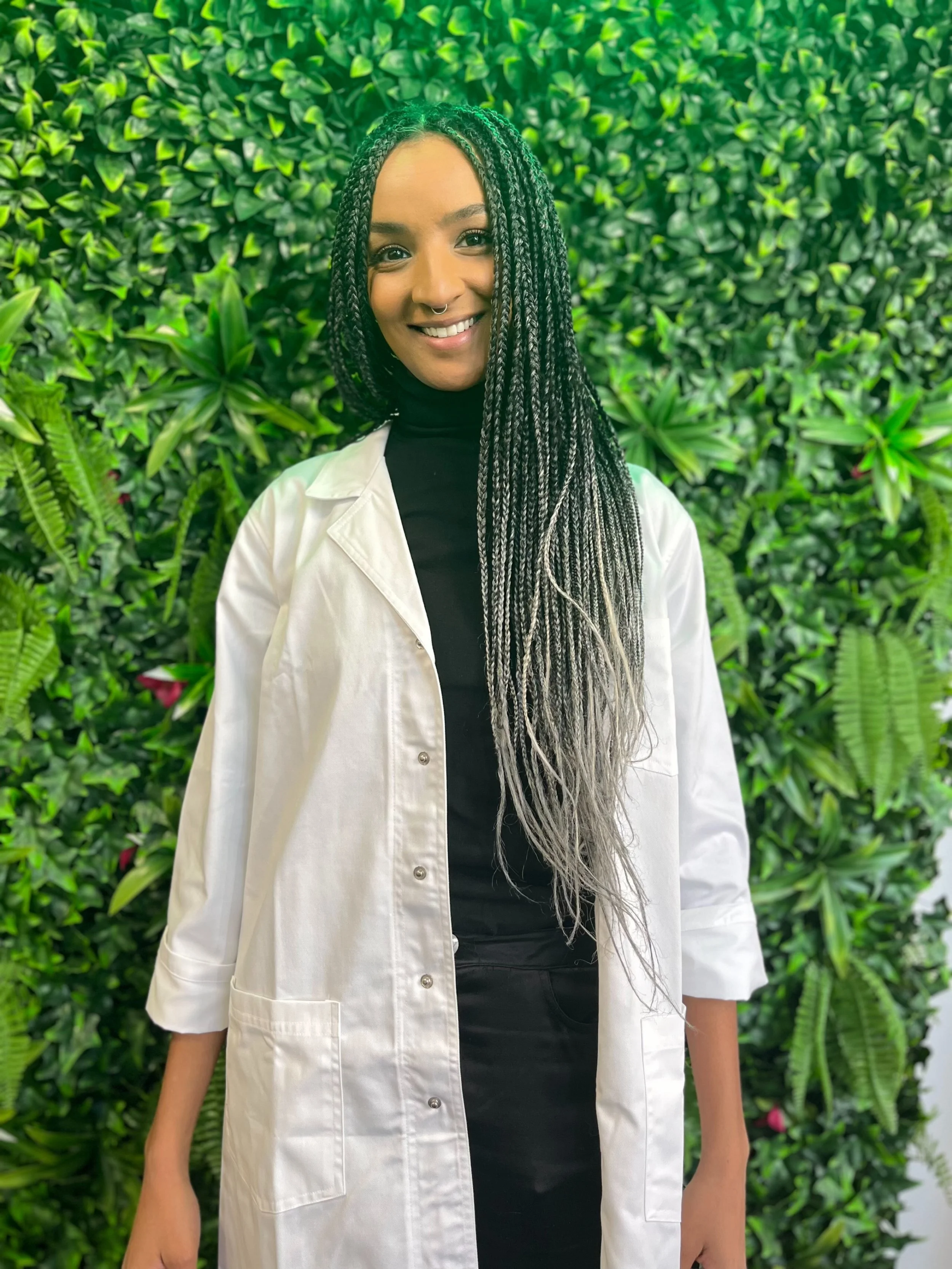 A young woman with long braided hair, smiling, wearing a black turtleneck and white coat, standing in front of a green leafy background.