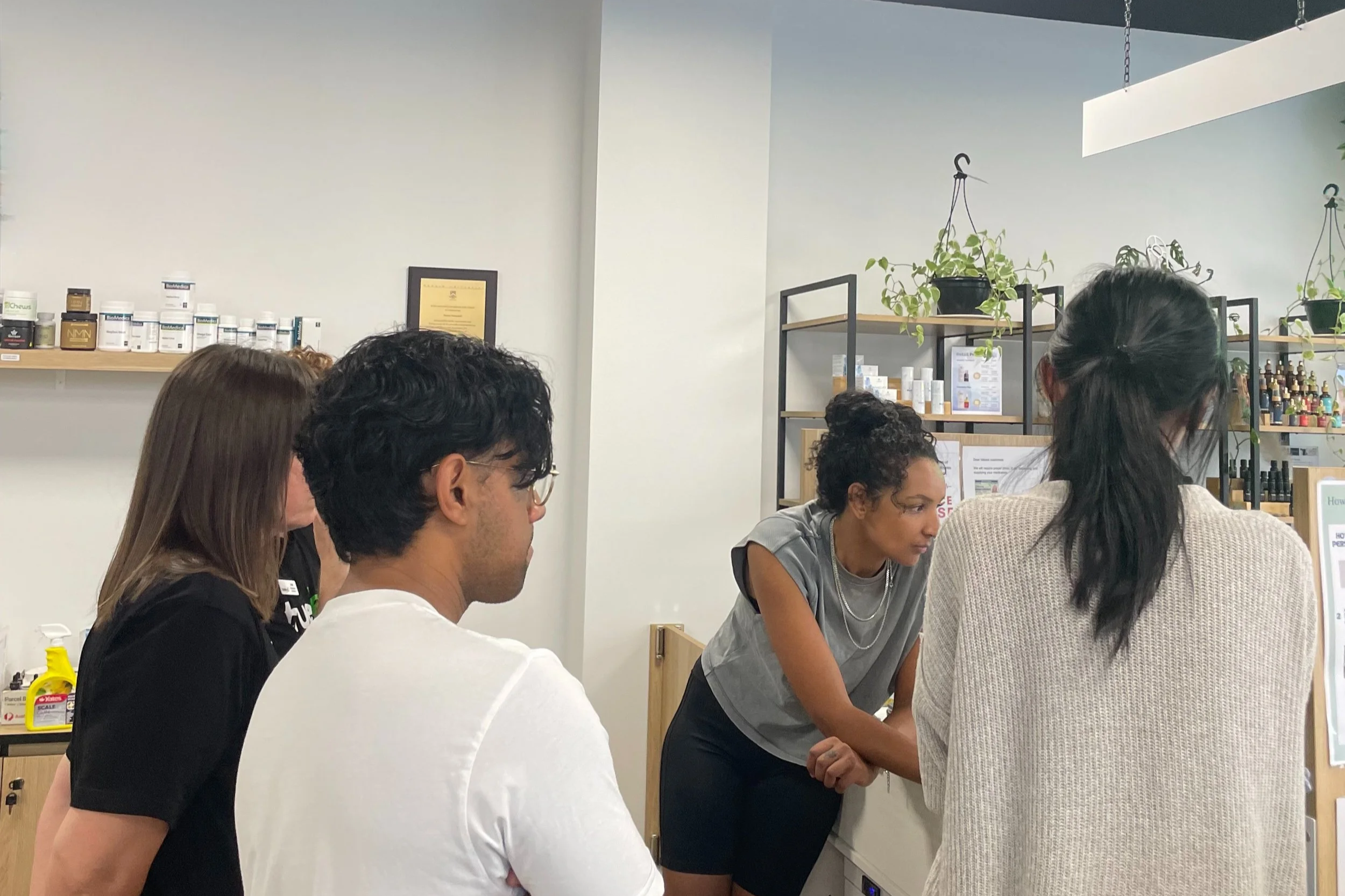 A group of diverse people sitting and standing around a table in a modern office or classroom, engaged in discussion or activity, with shelves of bottles and plants in the background.