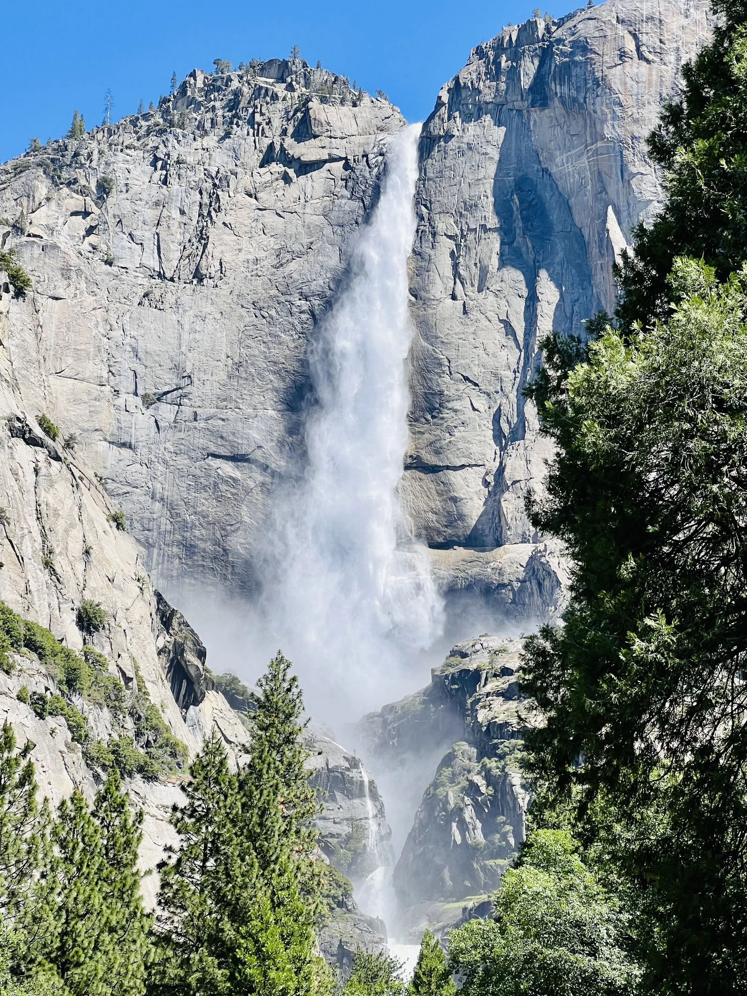 A tall waterfall cascading down the rocky cliffs of a mountain surrounded by green trees under a clear blue sky.