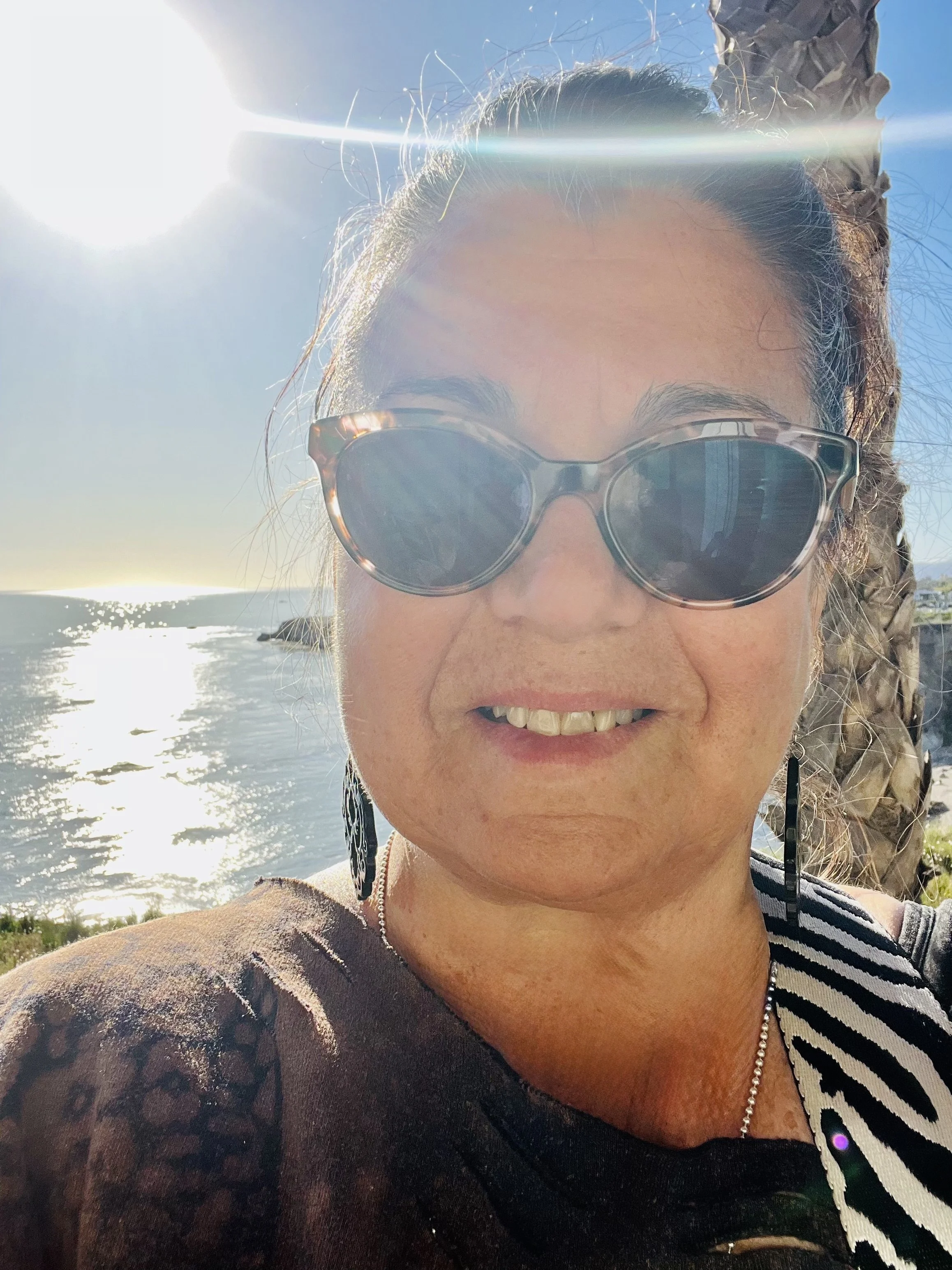 A woman wearing sunglasses and a striped shirt takes a selfie outdoors near the water during daytime, with the sun shining brightly in the sky.
