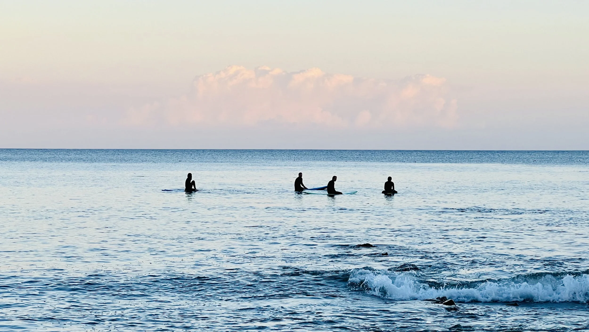 Four people silhouetted on surfboards sit in calm ocean water during sunset, with a pastel-colored sky and a few clouds in the background.
