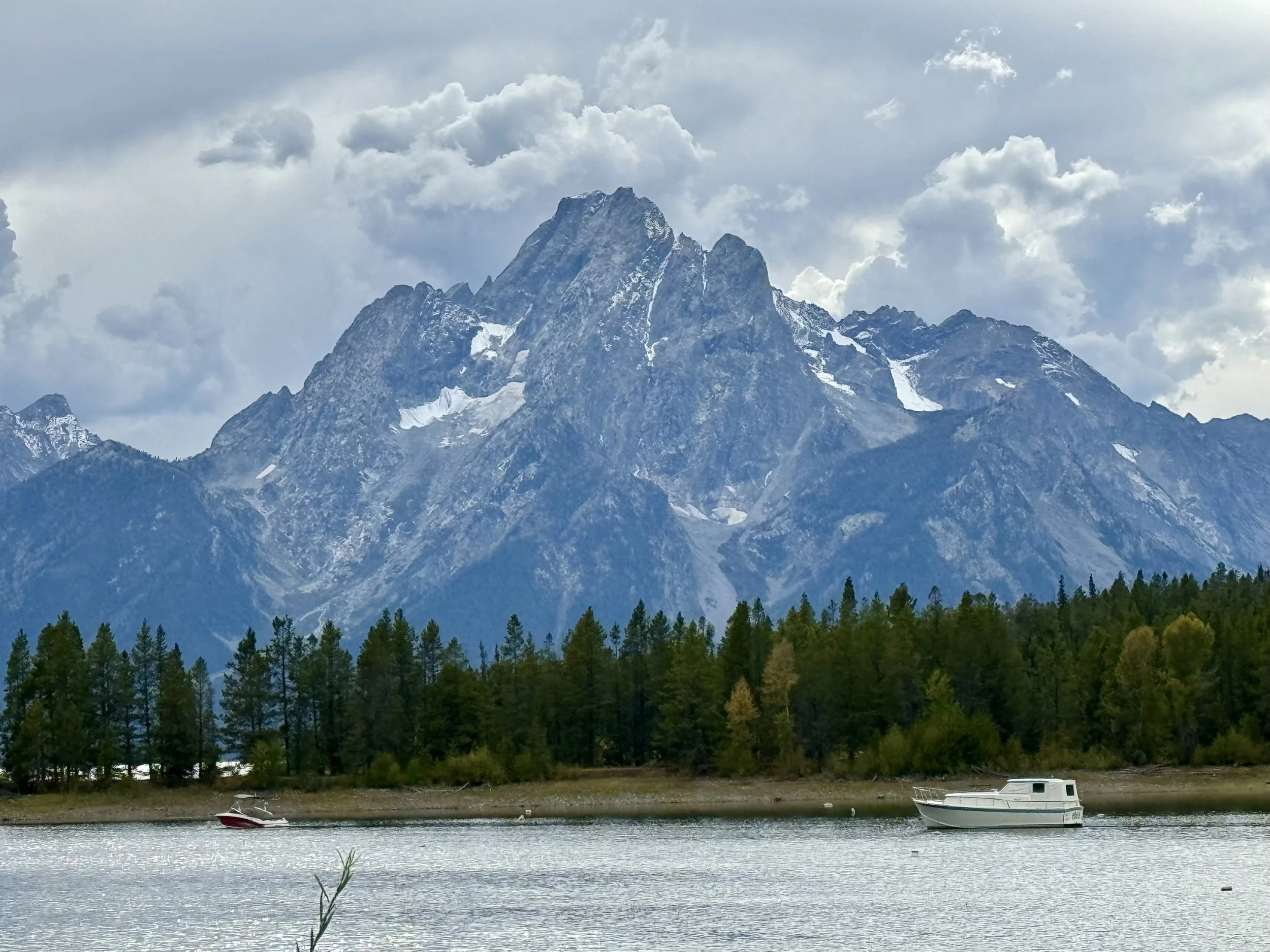 A mountain with snow patches rises behind a forest of green trees, near a body of water where two boats are floating, one red and one white.