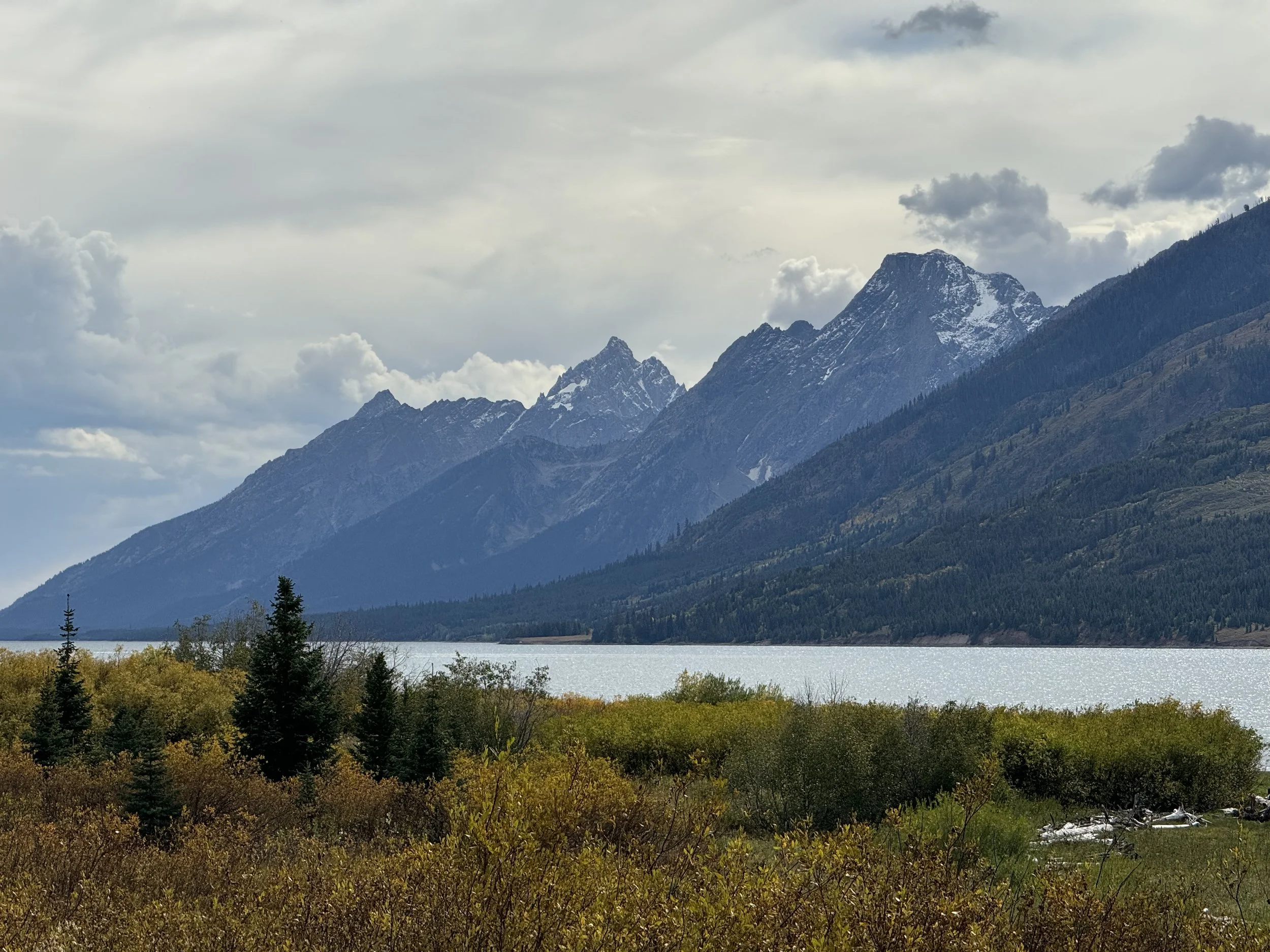 Scenic view of a mountain range with snow-capped peaks, a lake in the foreground, and trees with autumn foliage.