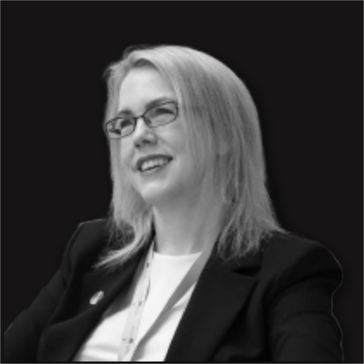 Black and white photo of a woman with glasses, shoulder-length hair, smiling, wearing a blazer and a lanyard.