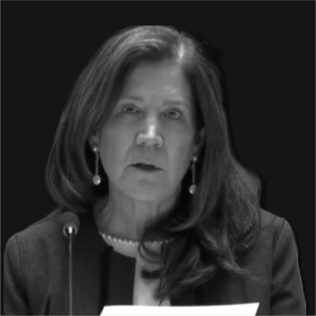 Black and white photo of a woman with medium-length hair, wearing earrings, a pearl necklace, and a blazer, speaking or reading at a podium.
