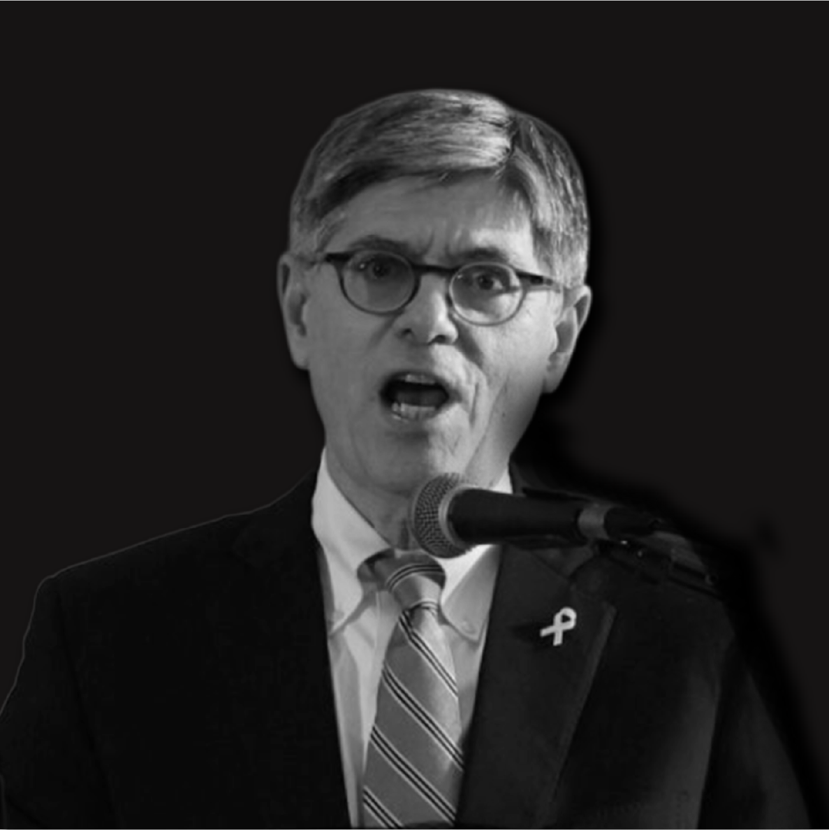 Black and white photograph of a man with glasses and gray hair speaking into a microphone, wearing a dark suit, white shirt, and striped tie, with a ribbon lapel pin.