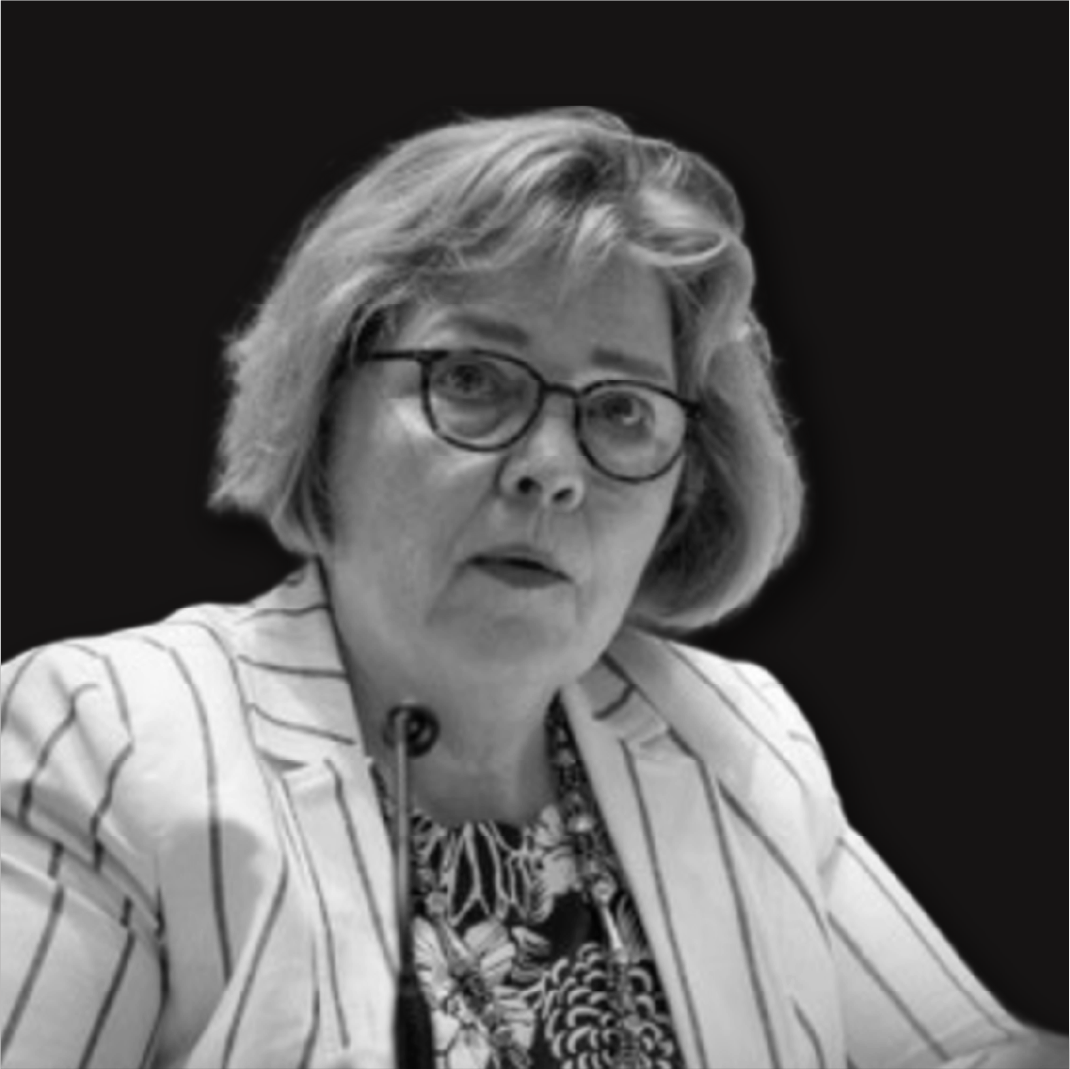 Black and white photo of a woman with glasses and short hair, wearing a striped blazer and a patterned blouse, speaking into a microphone.