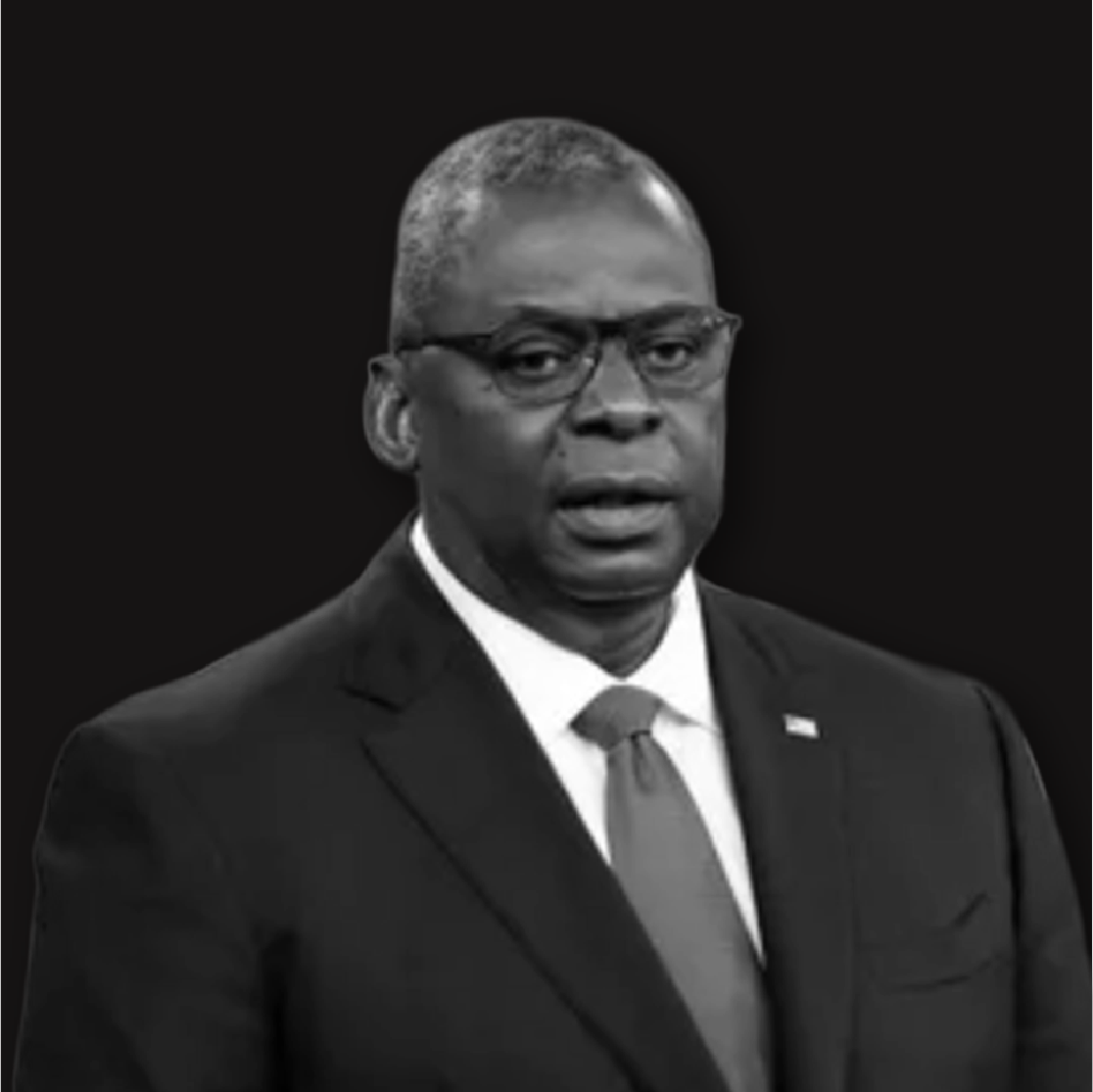 Black and white photo of a man wearing glasses, a suit, and a tie, against a dark background.