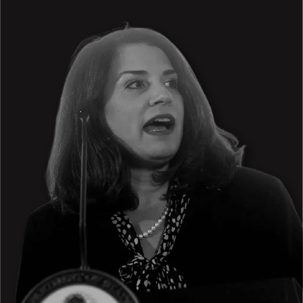 Black and white photo of a woman speaking at a podium, wearing a dark blazer, a polka dot blouse, and a pearl necklace.