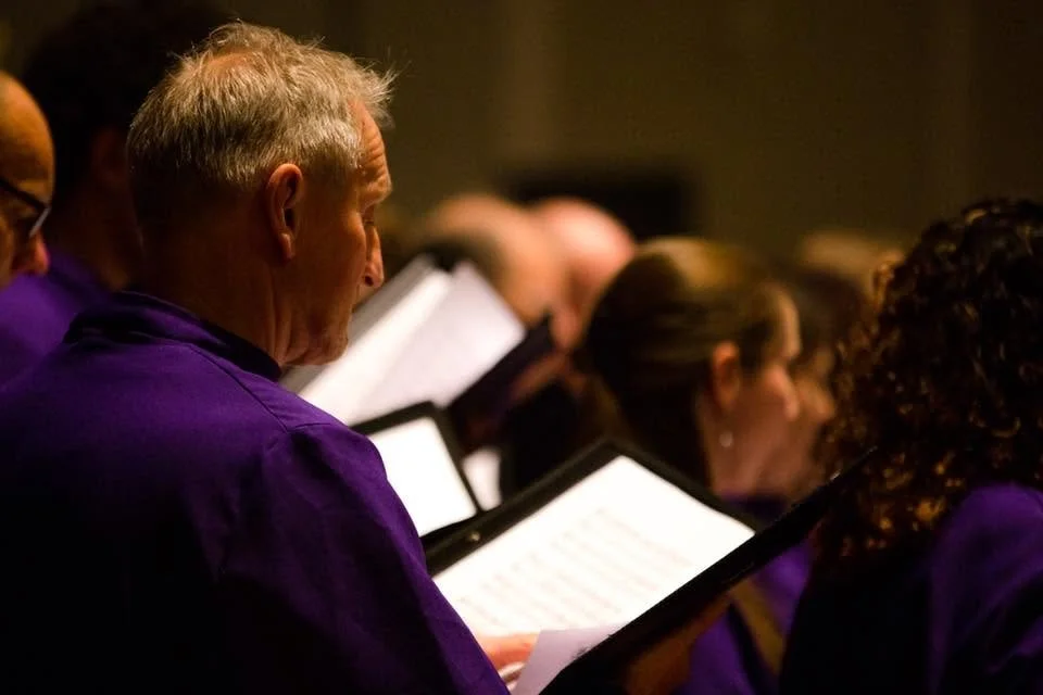 Group of choir singers dressed in purple robes, reading music sheets during a performance.