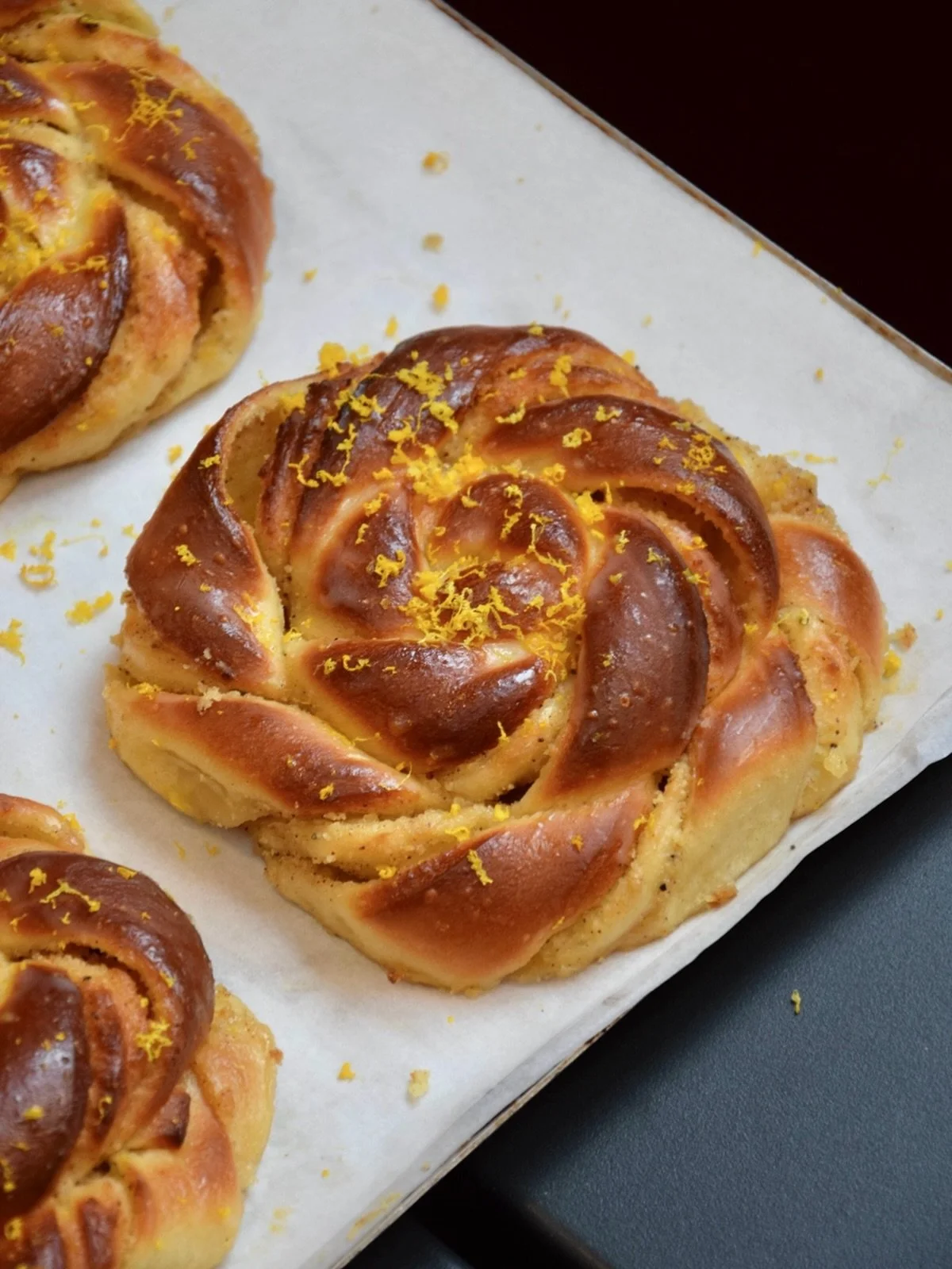 Bakery pastries with a rosette or flower shape, topped with lemon zest, on a white parchment-lined tray.