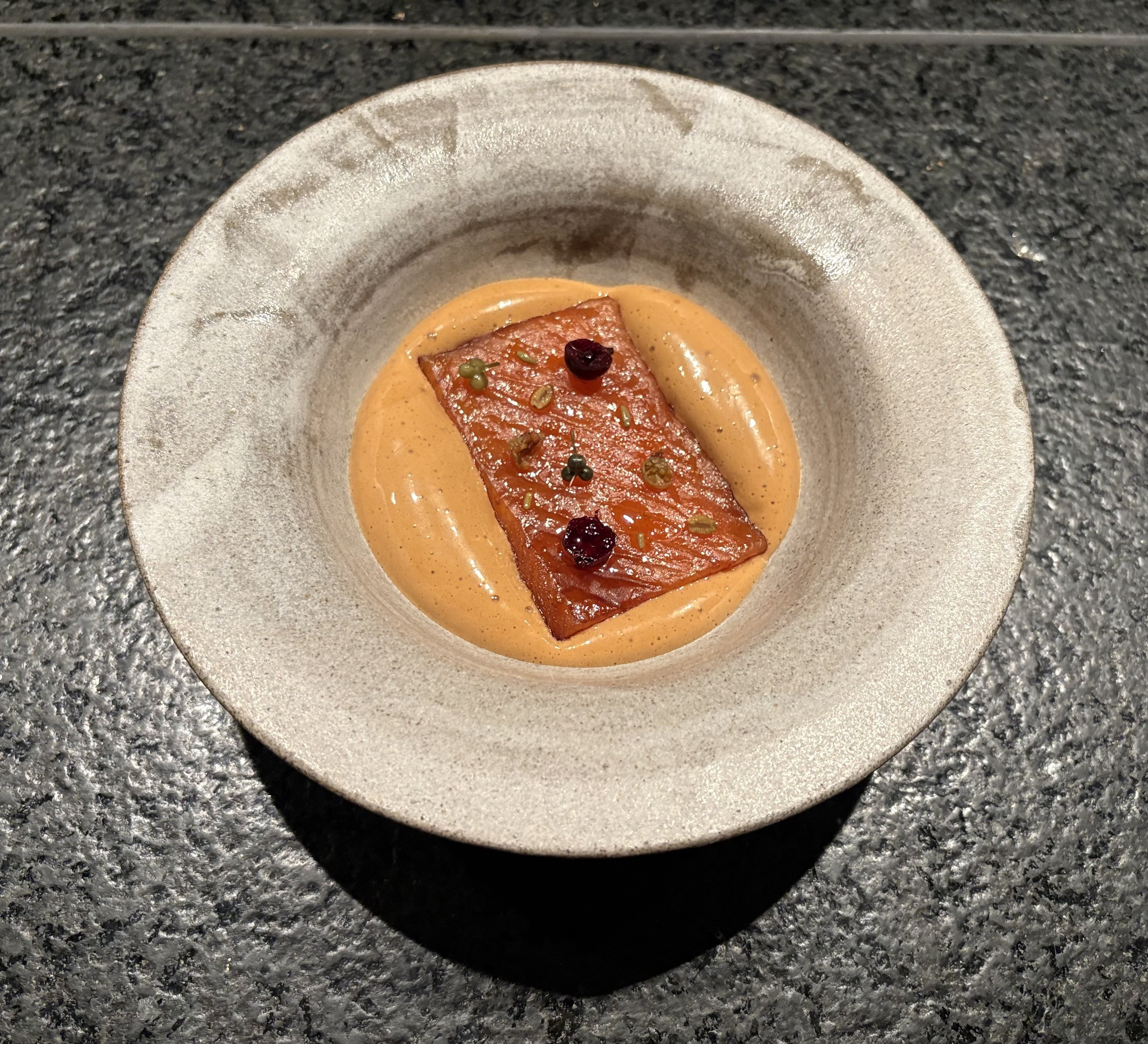 Close-up of a gourmet appetizer on a beige plate with textured surface, featuring a piece of smoked salmon garnished with capers and a dark berry or pepper, on a black speckled countertop.