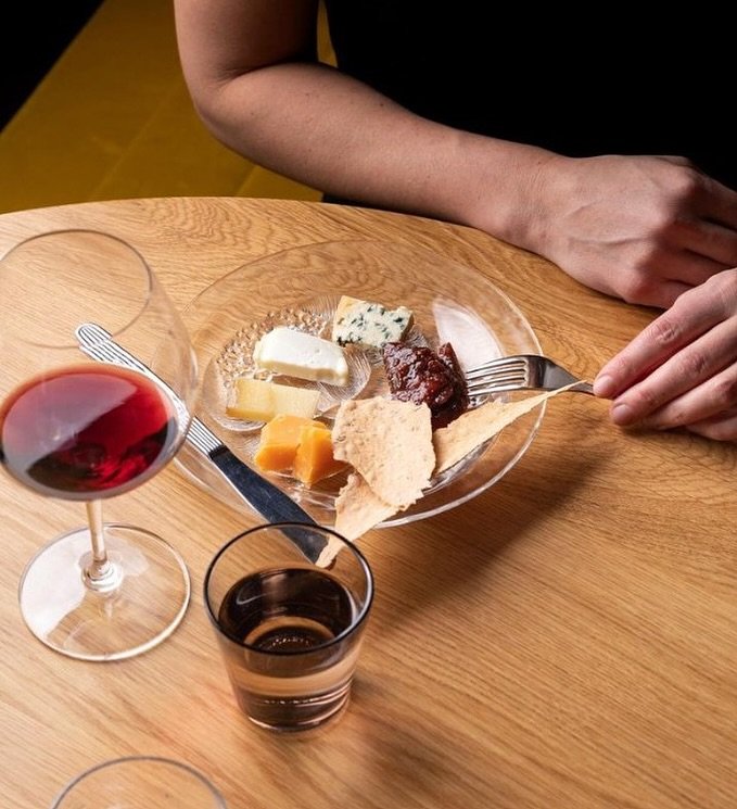 A person with fair skin is sitting at a wooden table with cheese and jam on a clear glass plate, a knife, and two glasses of red and dark beverages.