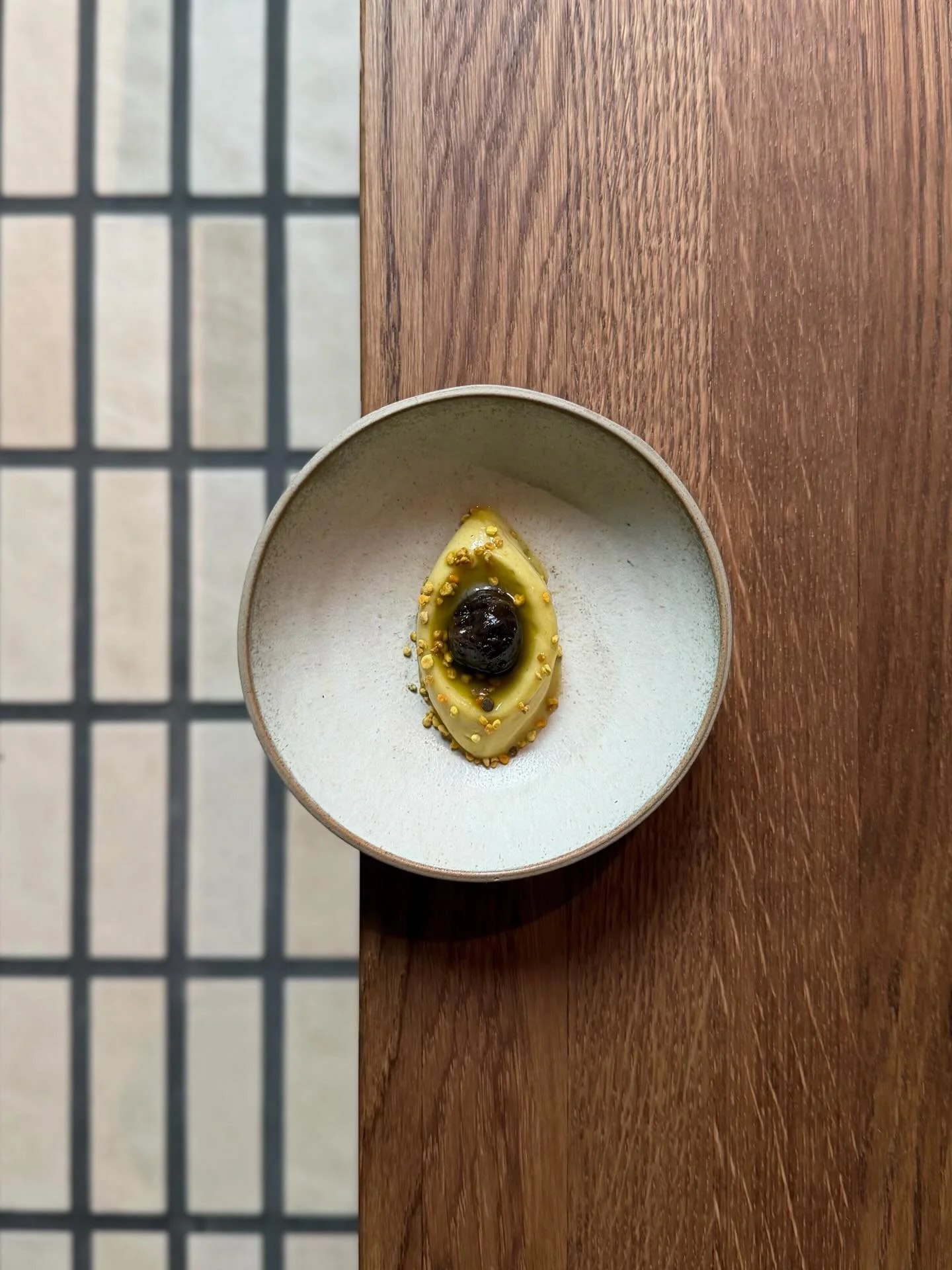 A small round dish with a yellow eye-shaped dessert topped with black and yellow garnishes, placed on a wooden surface.