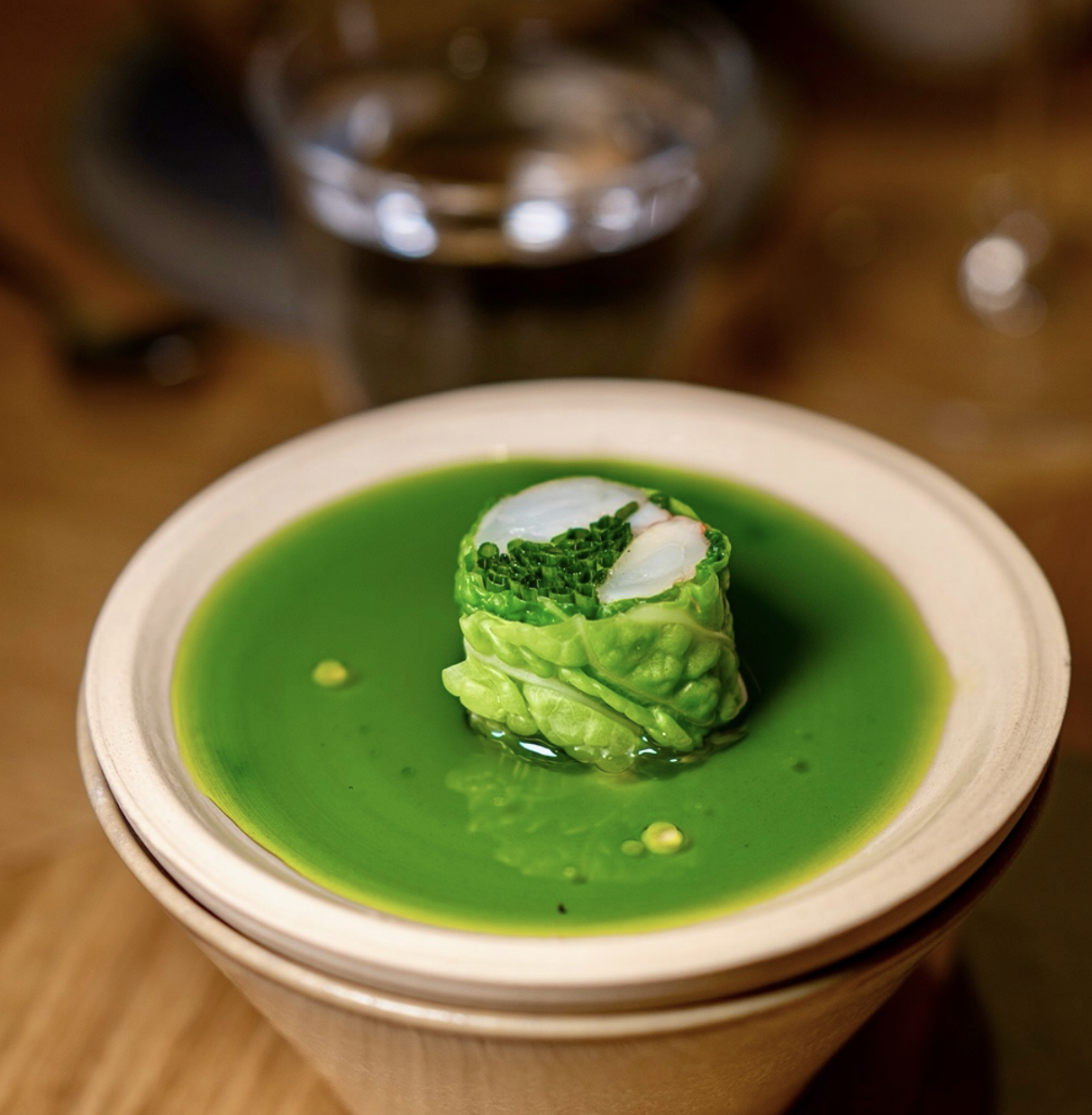 A wooden cup containing a vibrant green liquid with a rolled vegetable wrap inside, topped with chopped chives, placed on a wooden table.
