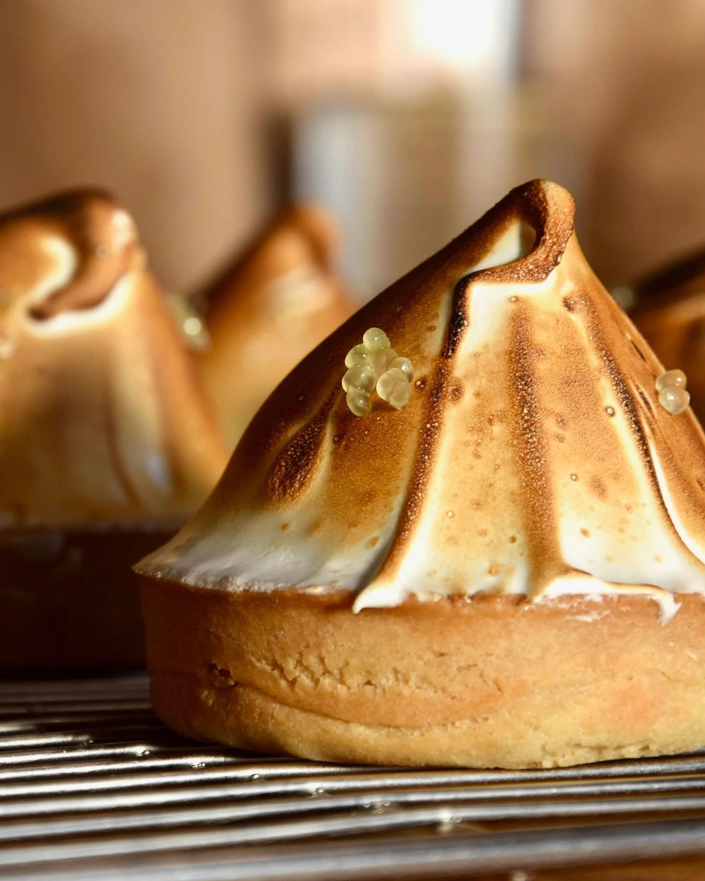 Close-up of a toasted meringue tart with a golden-brown peak, small sugar pearls on top, sitting on a cooling rack.