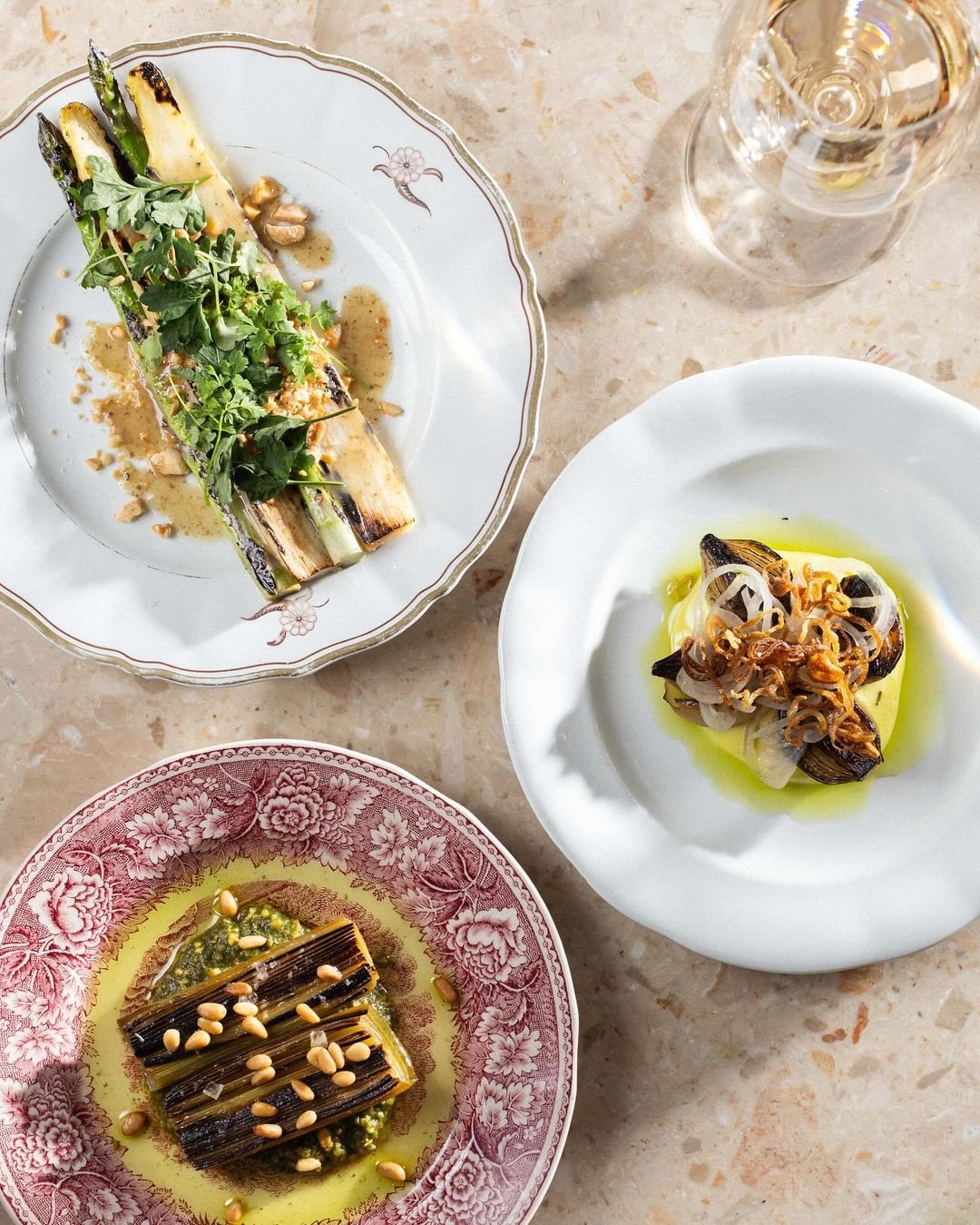 Three dishes of gourmet grilled vegetables topped with herbs, onions, and pine nuts on patterned porcelain plates, with a glass of white wine on a marble table.