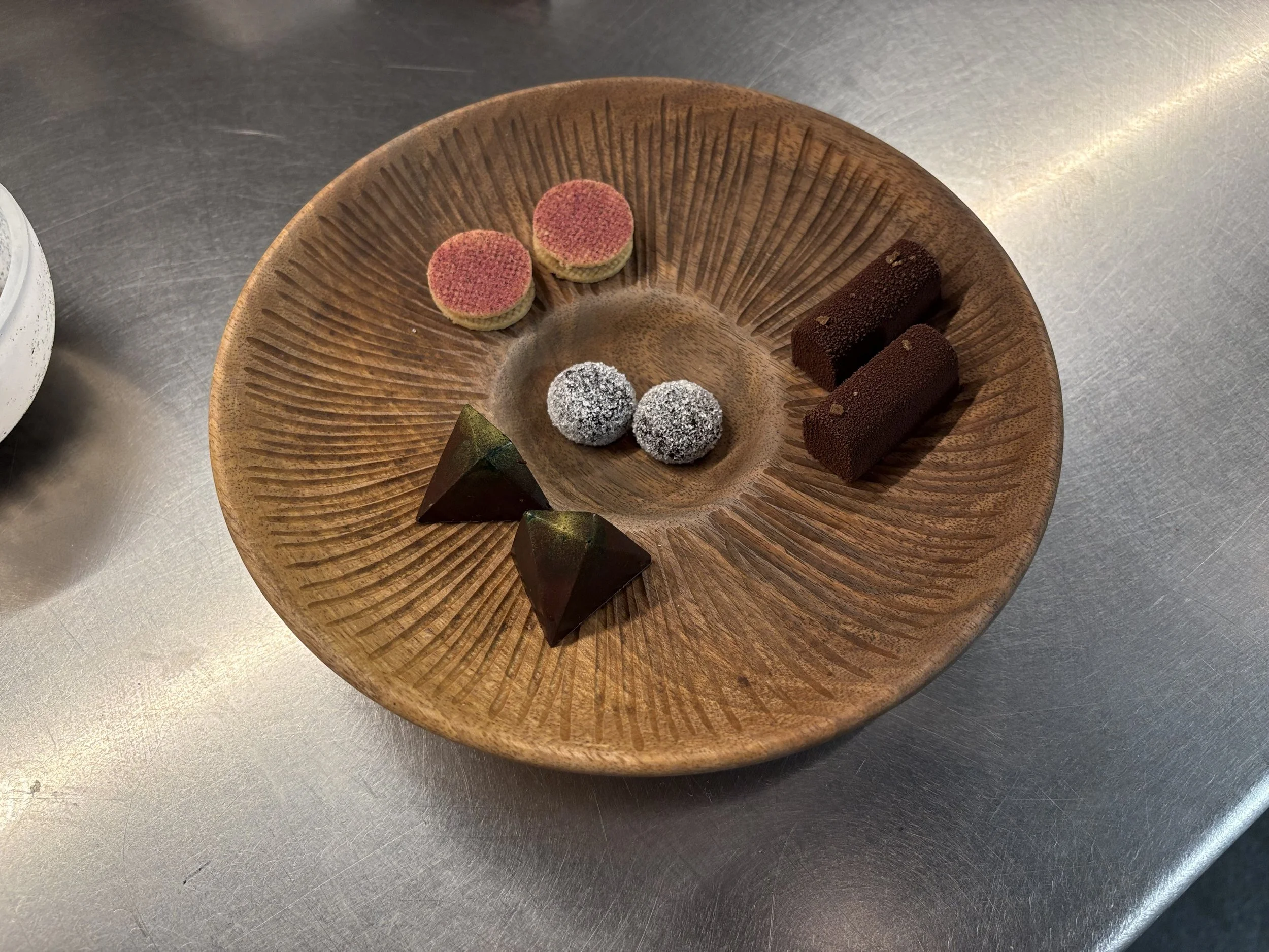 A round wooden plate with various small desserts including two pink and yellow round desserts, two silver-dusted spherical desserts, two brown rectangular desserts, and two dark triangular desserts.