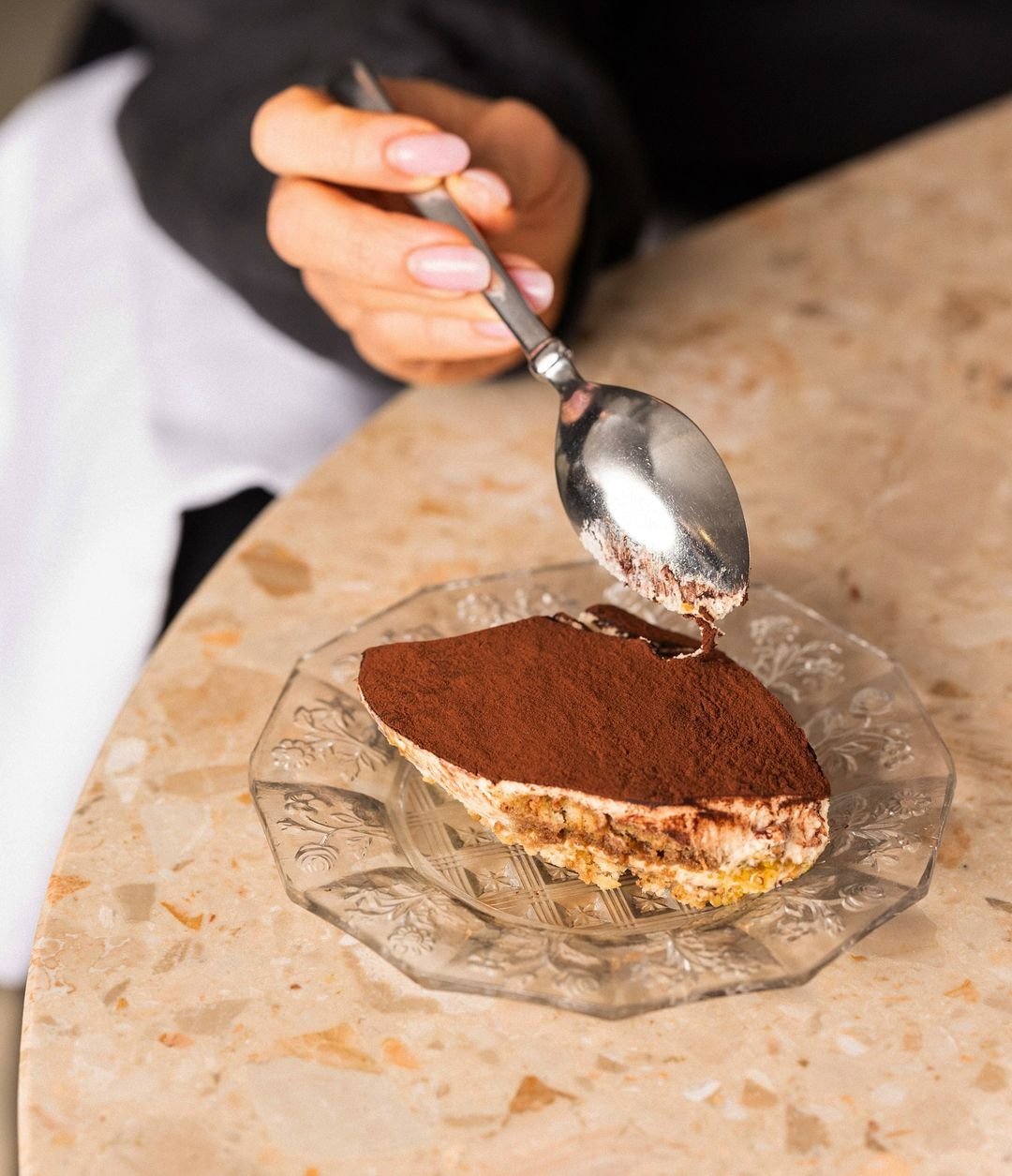 Person's hand holding a spoon over a slice of tiramisu cake on a glass plate.