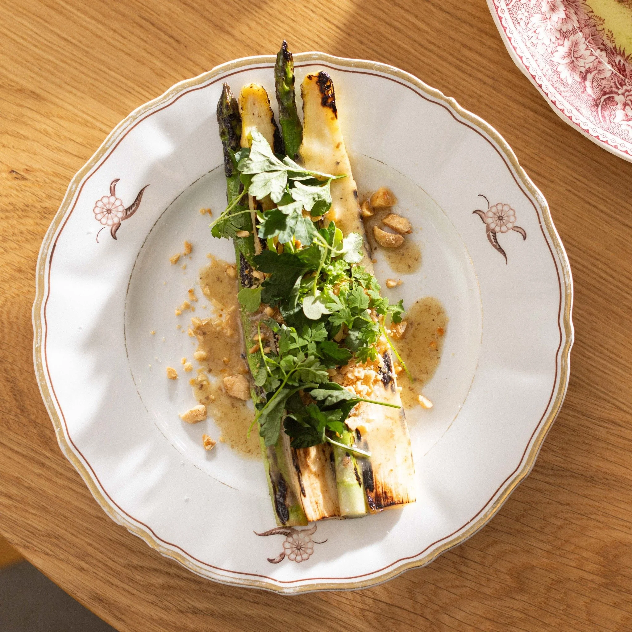 A white serving plate with gold trim and floral design, containing grilled asparagus topped with herbs and garnished with herbs and chopped nuts, placed on a wooden surface.