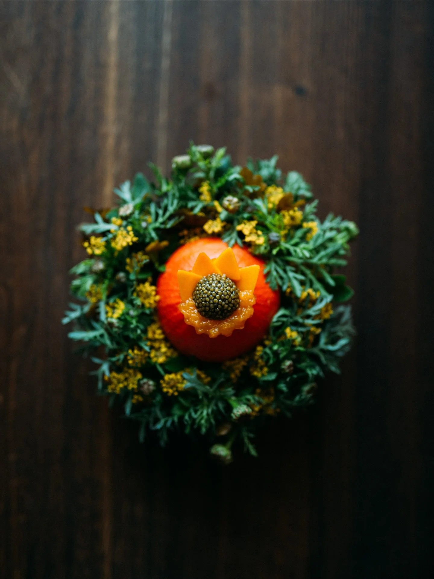 A pineapple decorated with mango slices and a blackberry, surrounded by green foliage and small yellow flowers, on a dark wooden surface.