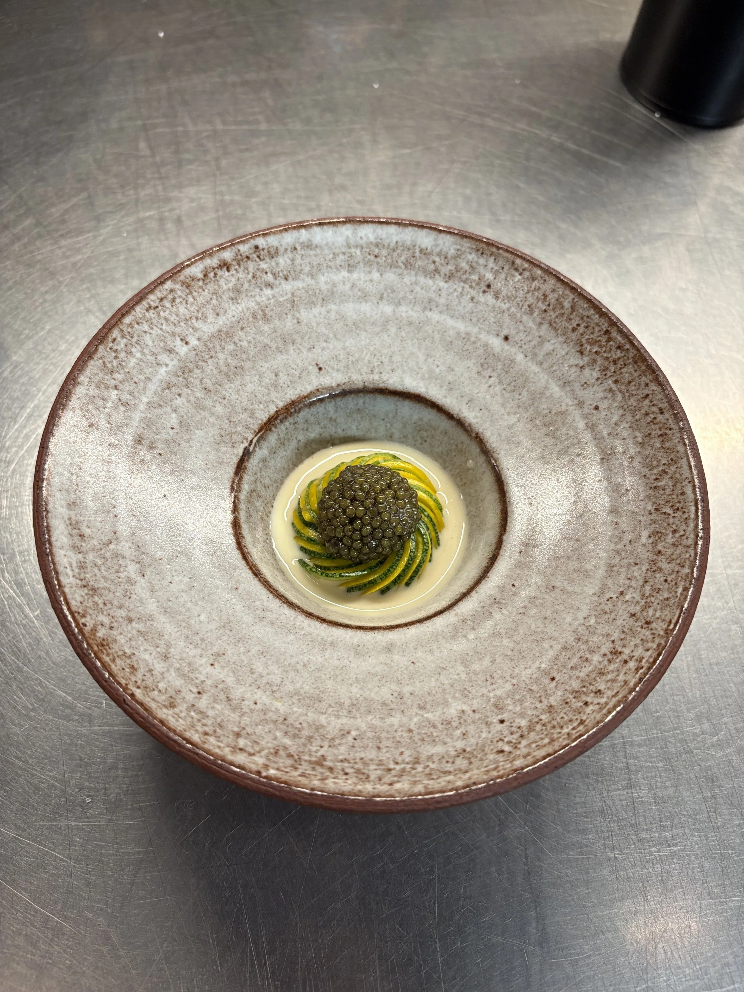 A ceramic bowl with a speckled brown and beige glaze, containing a dish with thinly sliced zucchini, topped with black caviar and surrounded by a creamy white sauce on a stainless steel countertop.