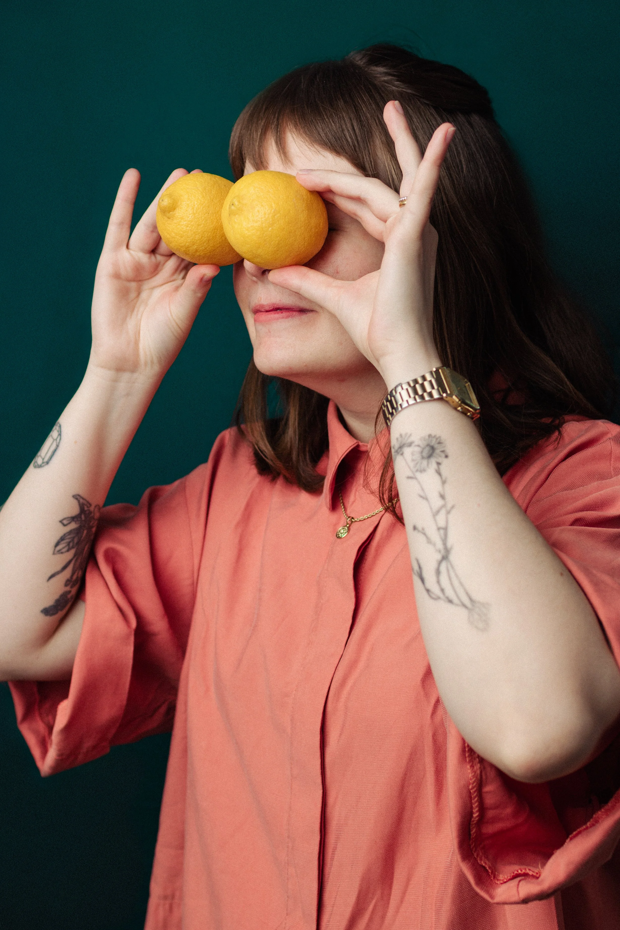 A woman with shoulder-length brown hair and tattoos on her arms is holding two yellow lemons in front of her eyes. She is wearing a copper-colored blouse, a gold watch, a delicate gold necklace, and a ring. The background is a dark teal color.