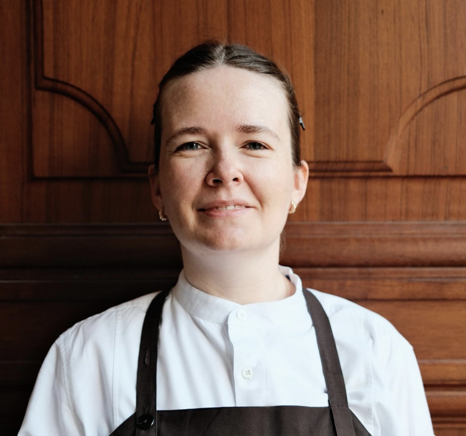 Close-up of a smiling woman with brown hair in a bun, wearing a white chef's coat and black apron, standing in front of a wooden background.