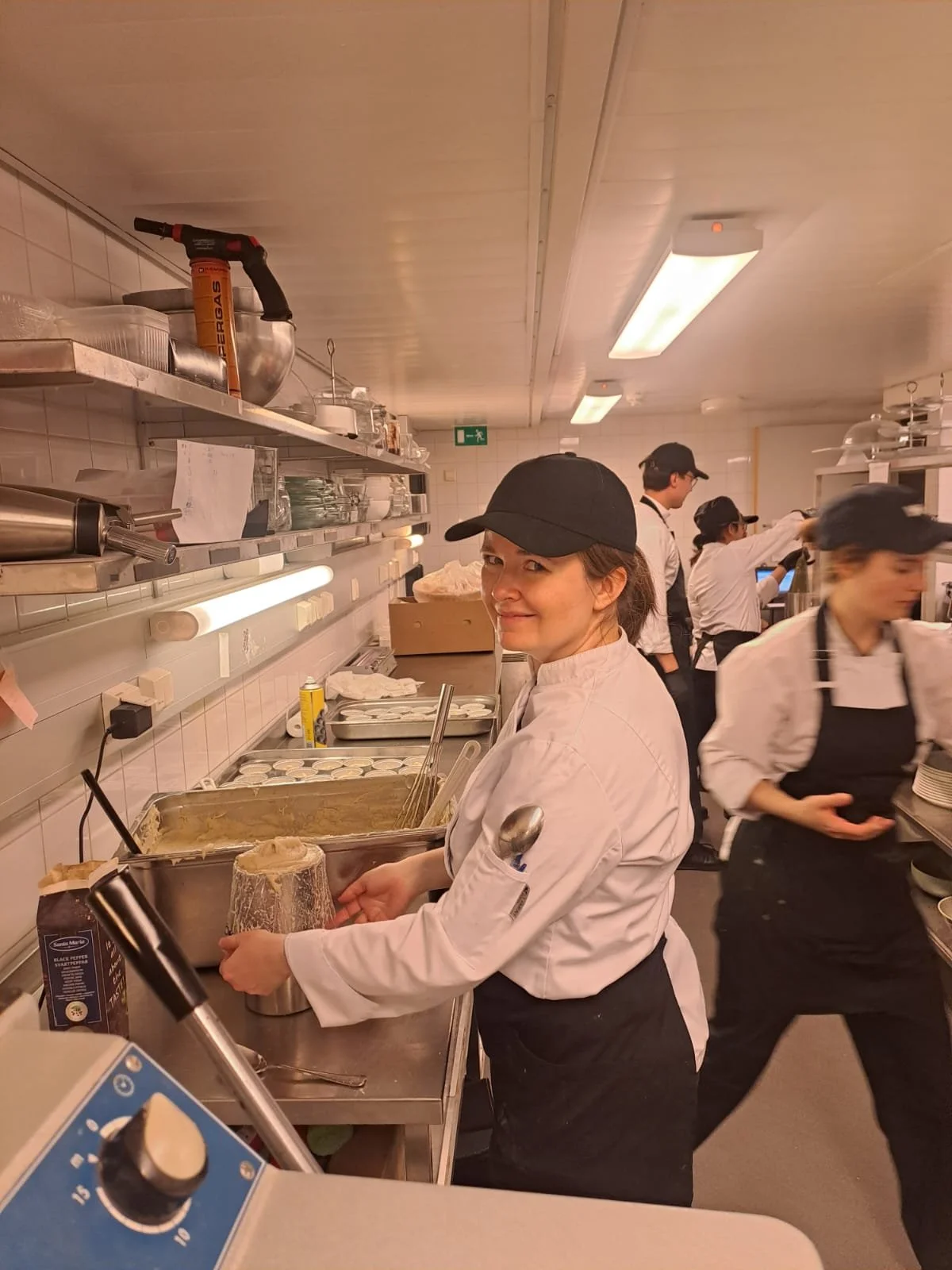 Kitchen staff preparing food, woman smiling at the camera, wearing a black cap and white chef coat.