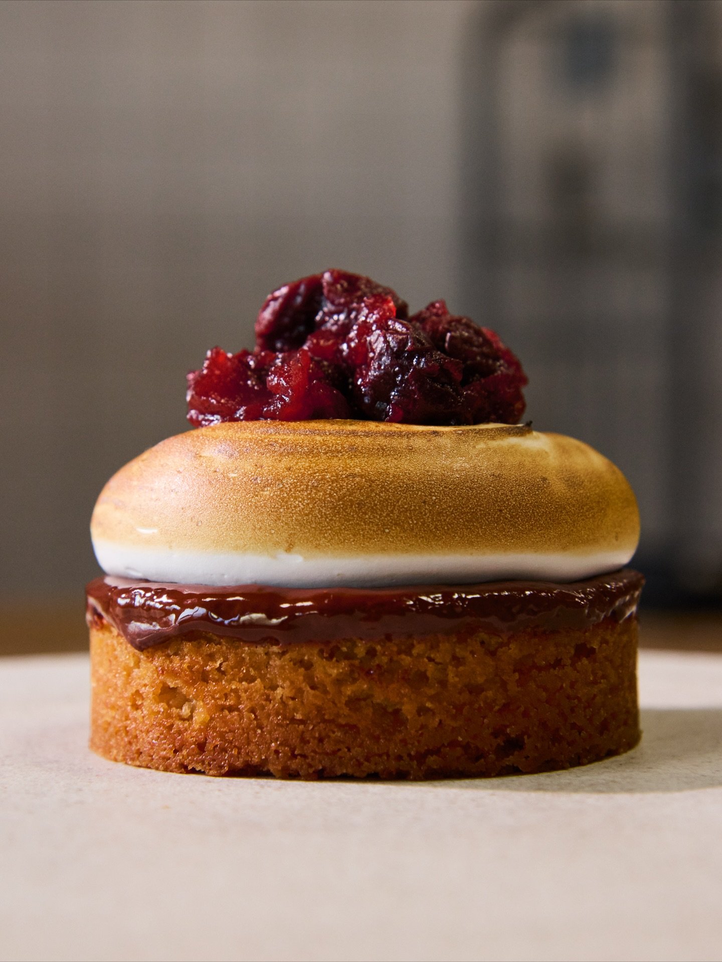 Close-up of a layered dessert with a shortbread cookie base, a layer of chocolate, a white cream layer, and a topping of cherry jam.