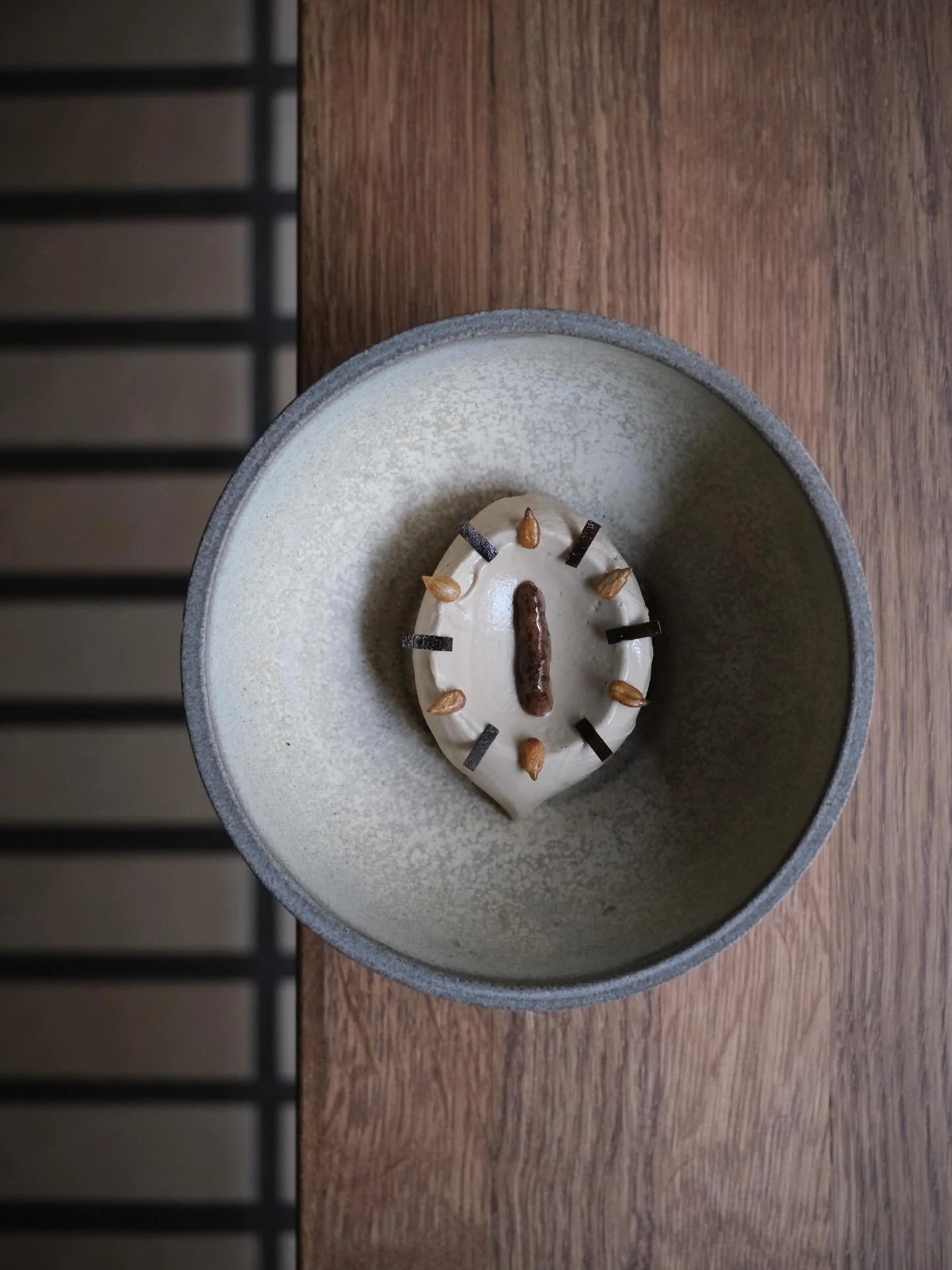 A round dessert cake with white frosting decorated with caramel drops and chocolate rectangles, in a ceramic bowl on a wooden surface.